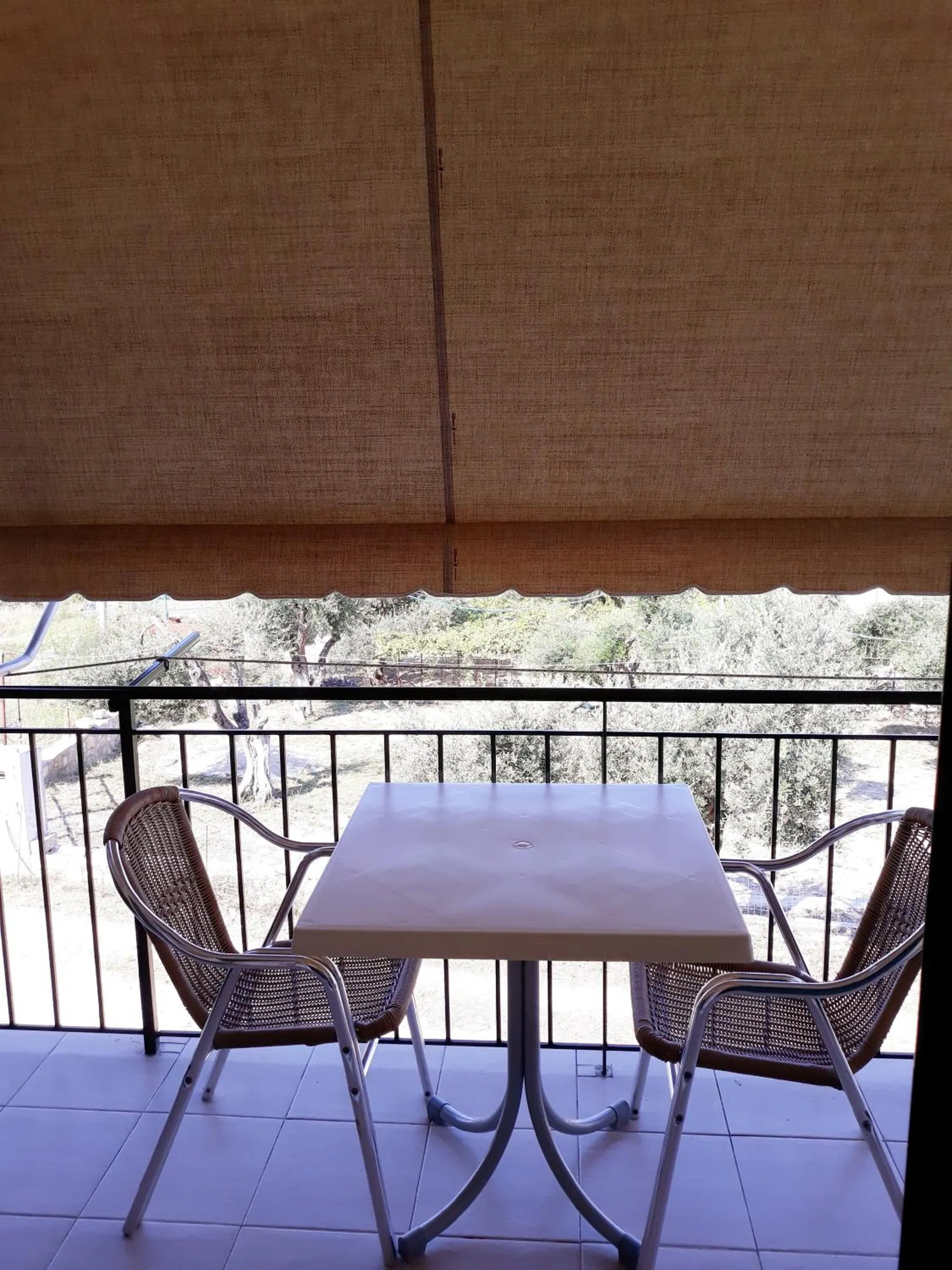 A balcony with a white table and two wicker chairs under a fabric awning, looking over the trees