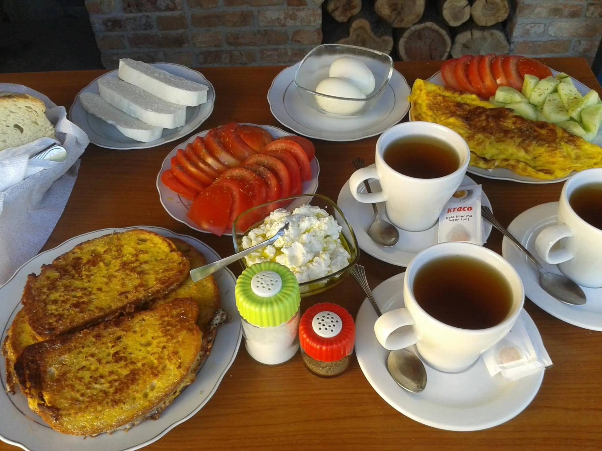 Breakfast on a wooden table: fried bread, an omelette, sliced tomatoes, white cheese, boiled eggs, cucumber and cups of tea