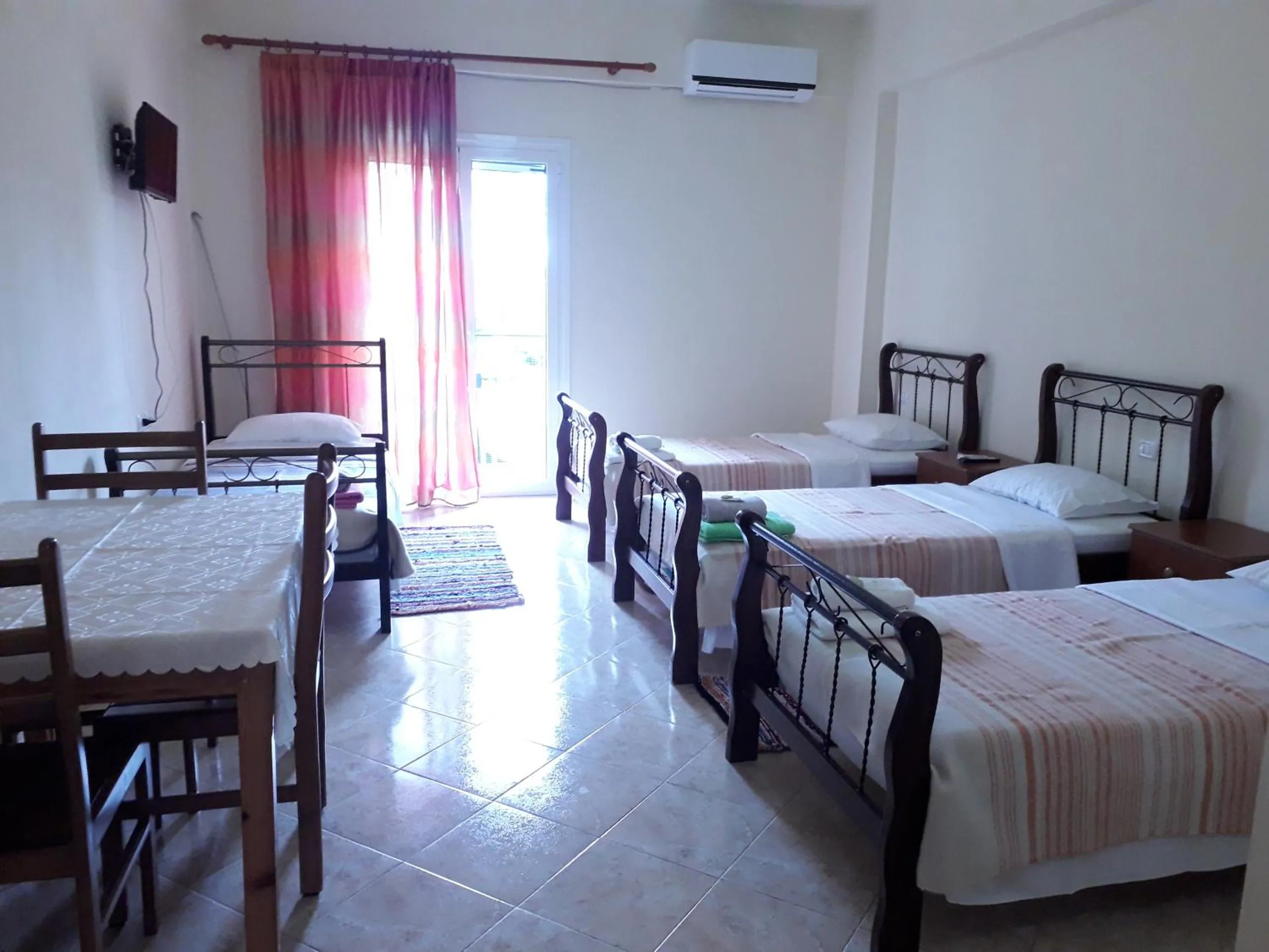 A quadruple room with four iron beds in a row, a dining table and a balcony door with pink curtains