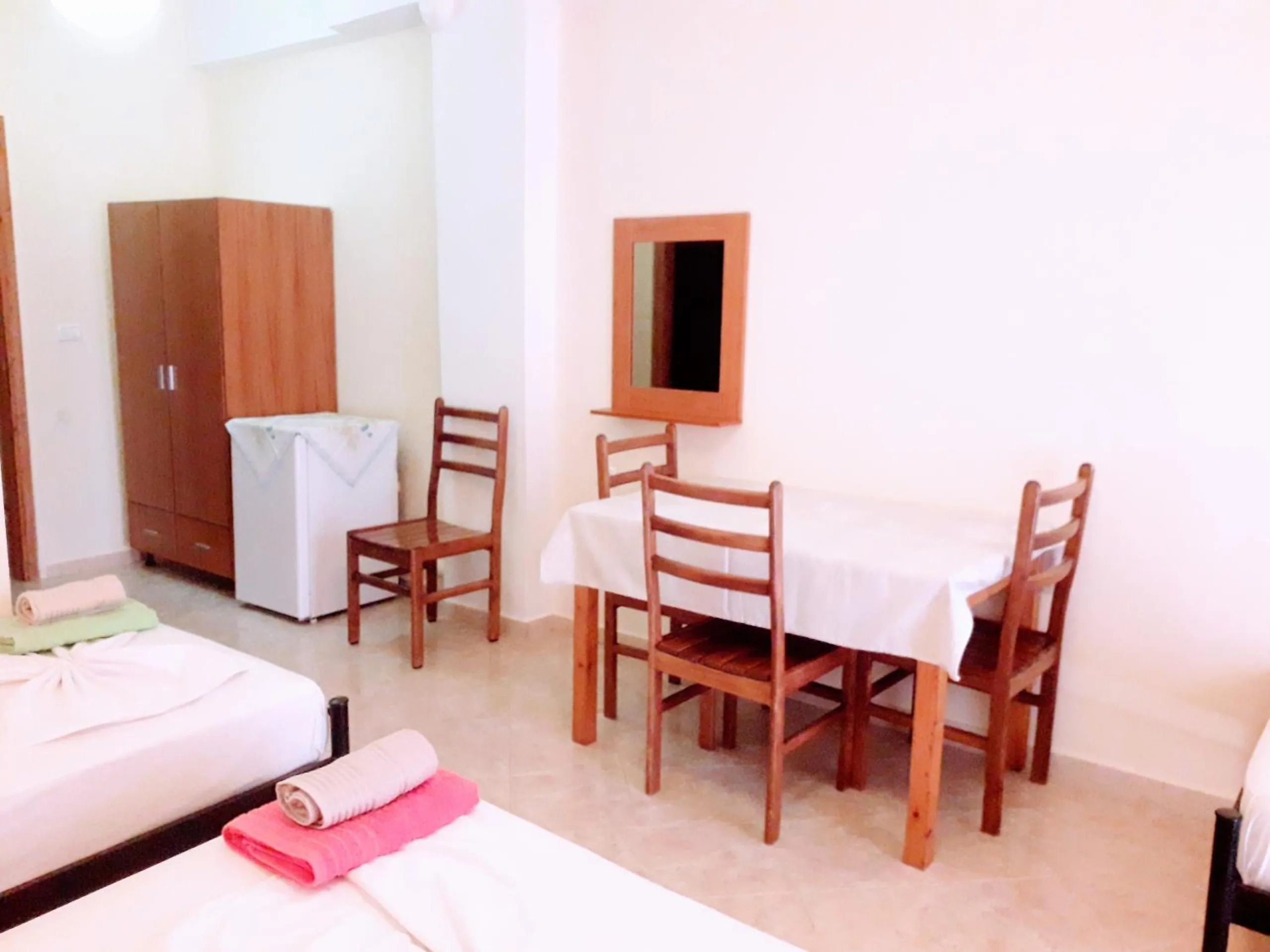 A room with a wooden wardrobe, a small refrigerator, a mirror and a dining table with four chairs