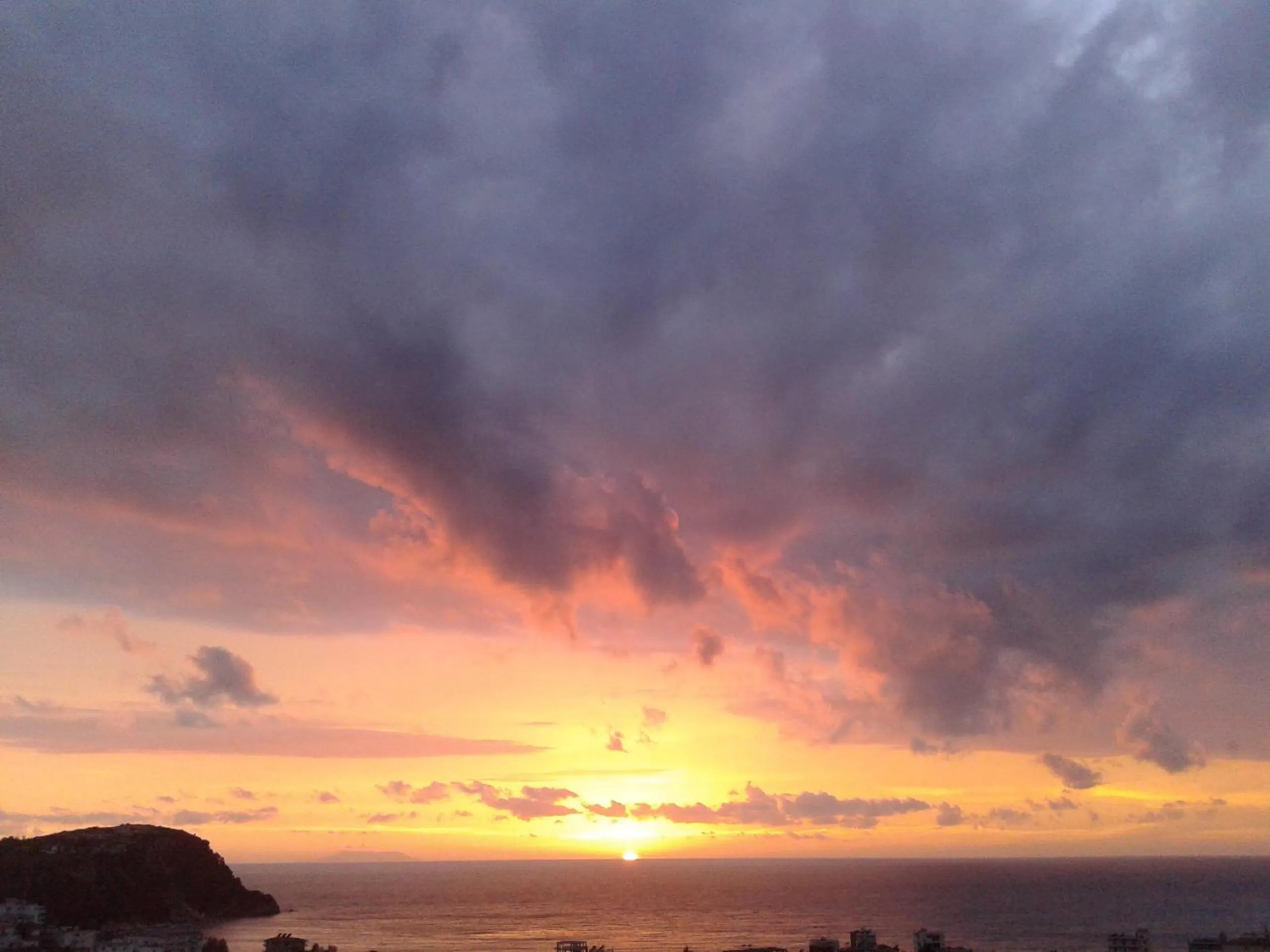 The sun setting into the sea beside a headland under a sky of dark cloud