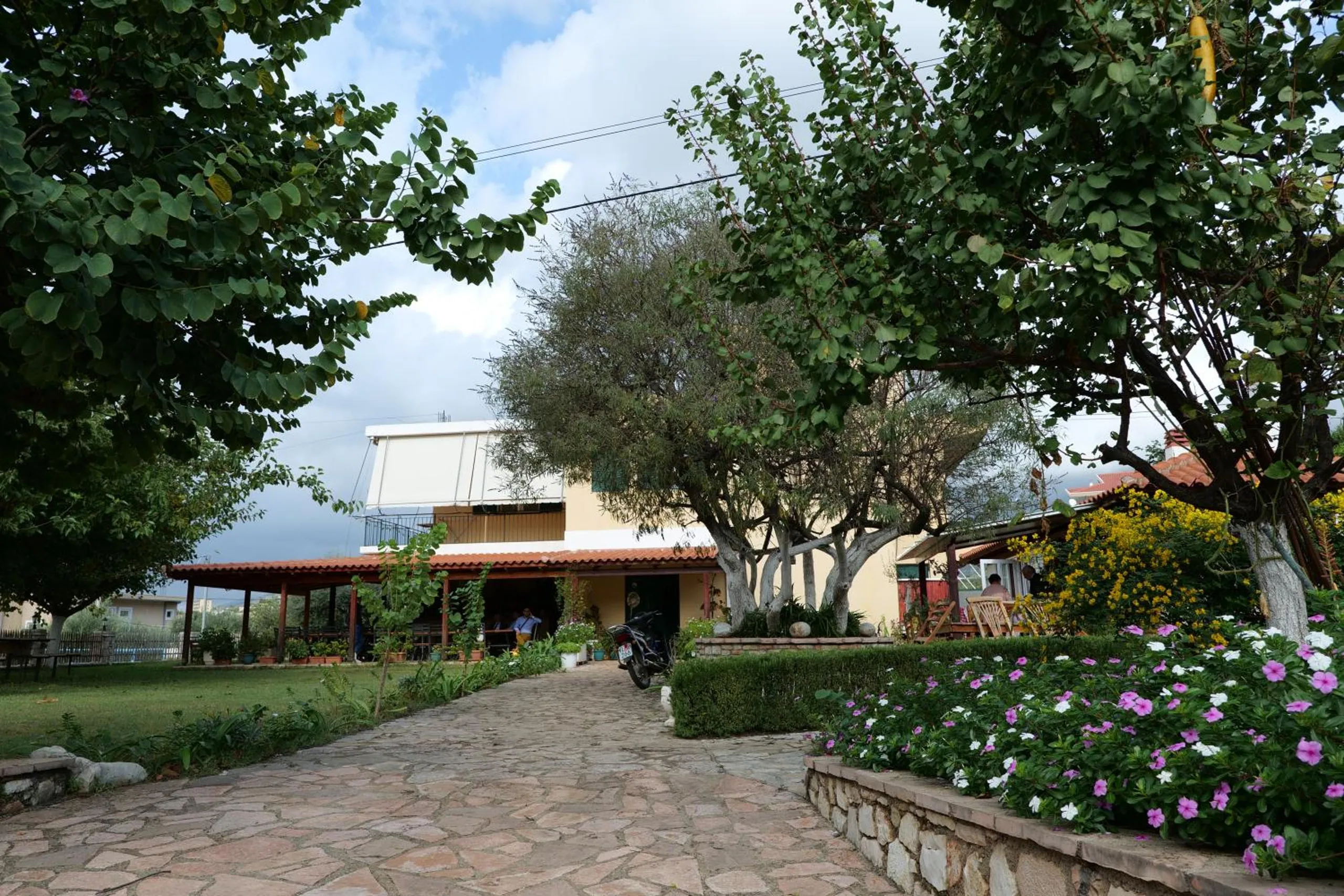 The cream two-storey house at Agrotourism Himara seen up a flagstone path between trees and beds of pink and white periwinkle
