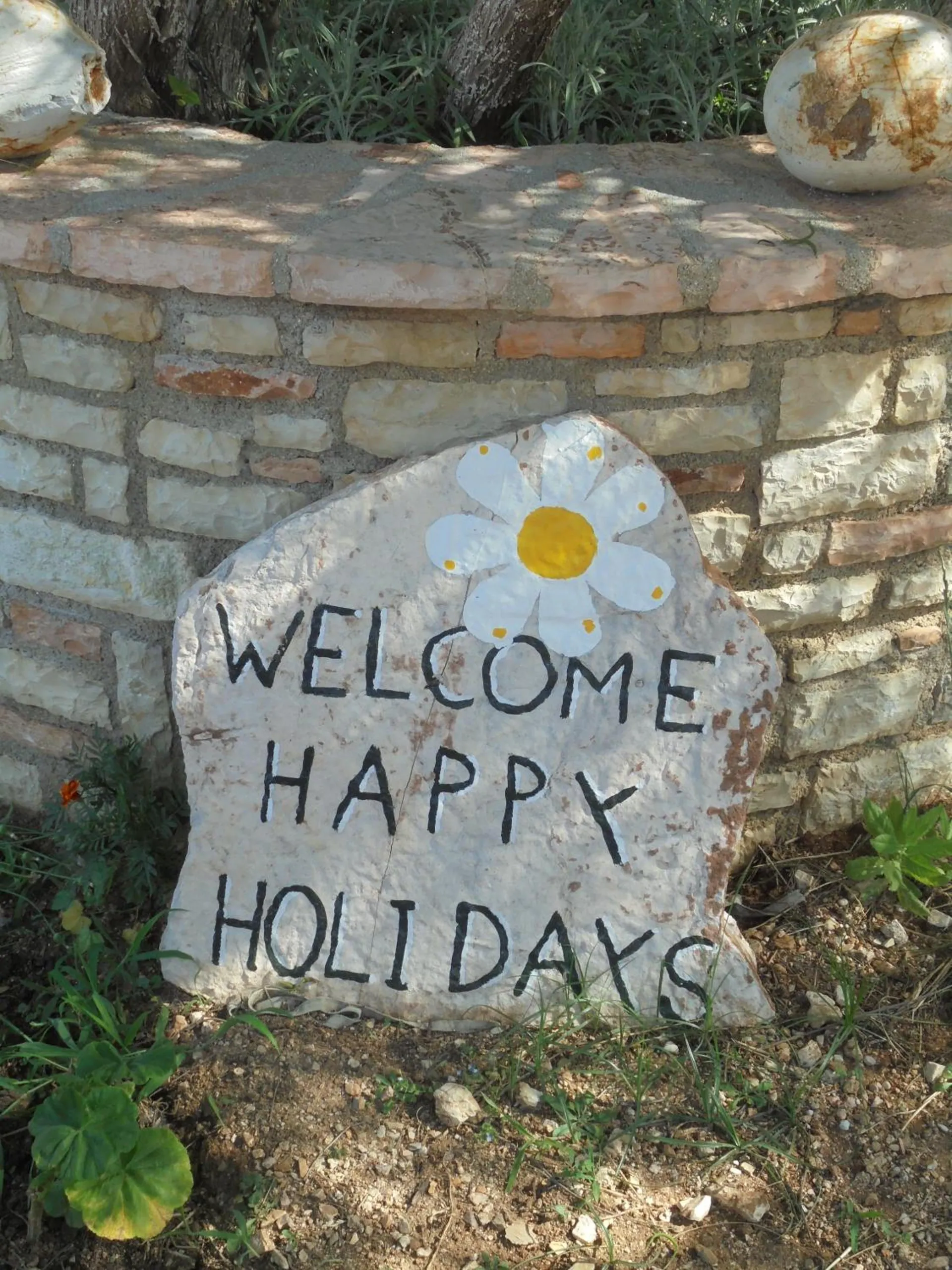 A white stone painted with a daisy and the words Welcome Happy Holidays, set against a stone wall