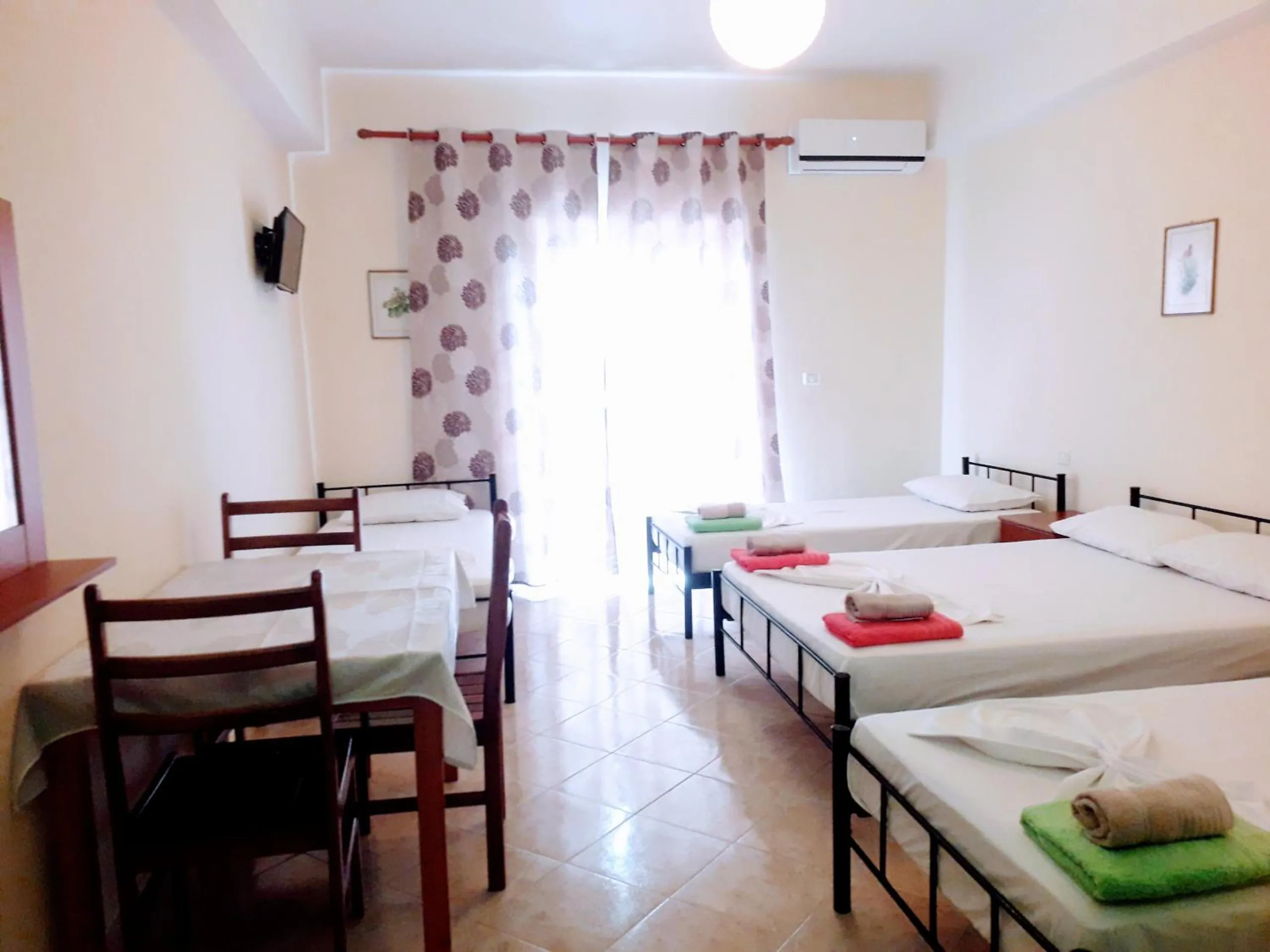 A bright triple room with three beds, a table and chairs, a wall-mounted television and a curtained balcony door