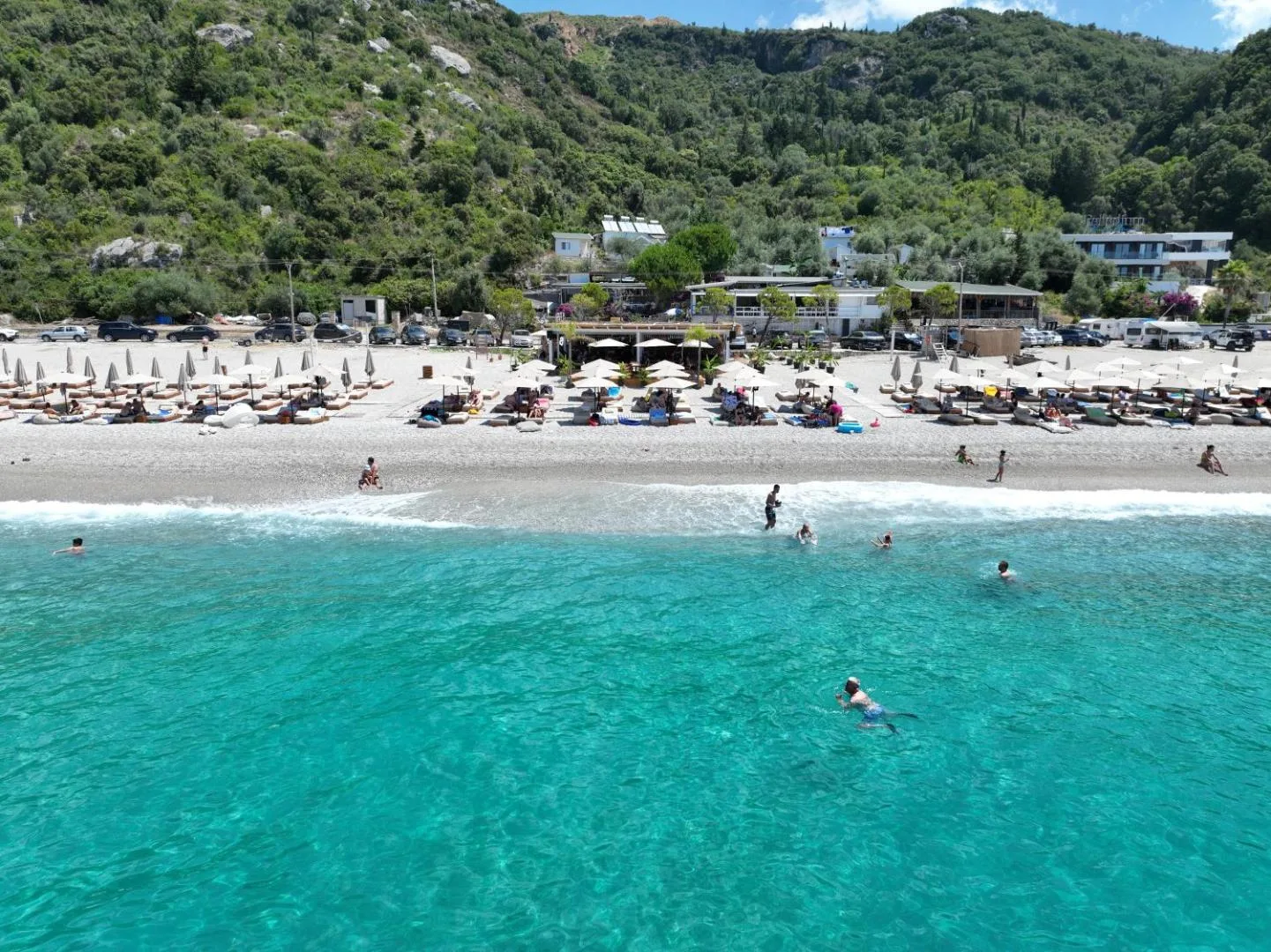 Swimmers in clear turquoise water in front of the loungers at the southern end of Dhërmi Beach, low buildings among the trees behind