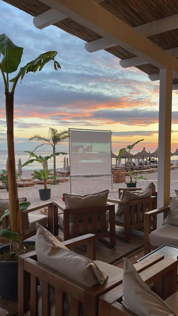 Timber sofas under the beach bar's reed roof at sunset, palms in pots and the sea glowing beyond