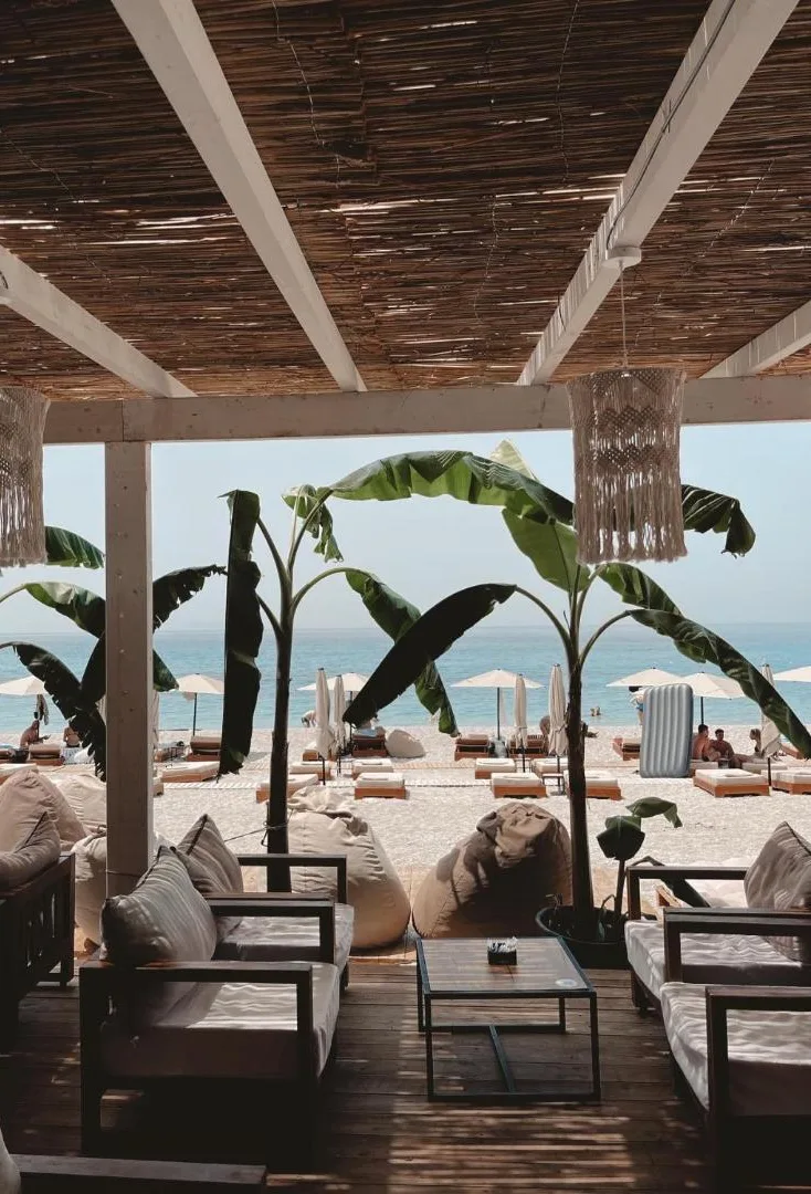 The beach bar lounge under a reed canopy with macramé lamps and banana plants, looking out to the umbrellas and the sea