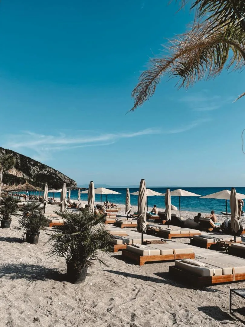 Cushioned wooden loungers and umbrellas on the beach under a palm, with the sea and the hills along the bay