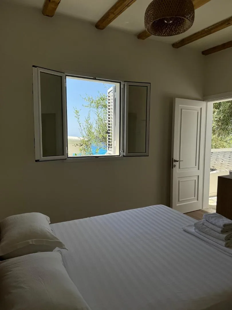 A beamed bedroom with its window shutters open to trees and a strip of sea