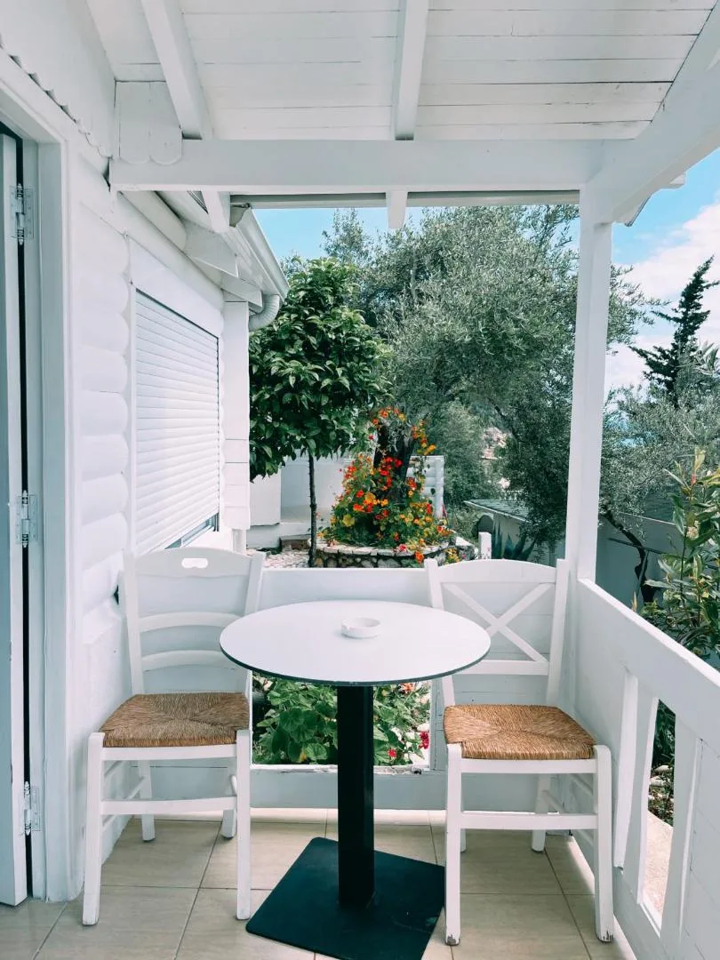 A bungalow veranda with a white rail, a round table and rush-seated chairs looking into olive trees