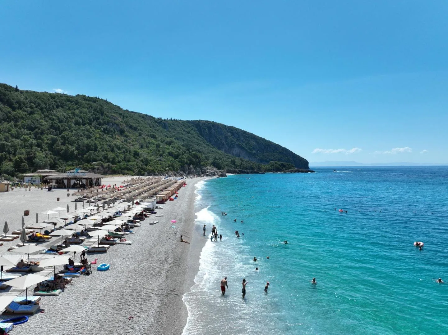 The pale shingle of Dhërmi Beach lined with umbrellas, running south to a wooded headland beside bright blue water