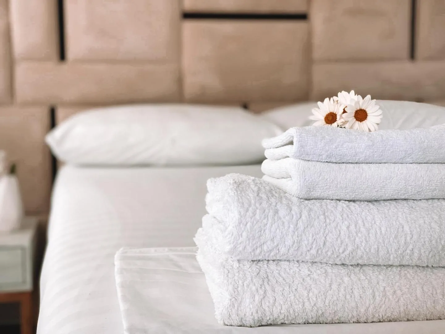 Folded white towels with a sprig of daisies on a made bed in front of a padded headboard