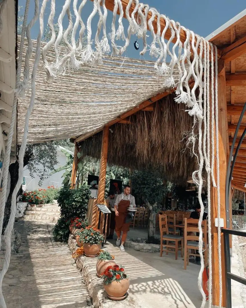 A stone path under a reed and rope pergola leading to the restaurant's thatched terrace, with potted geraniums along the wall