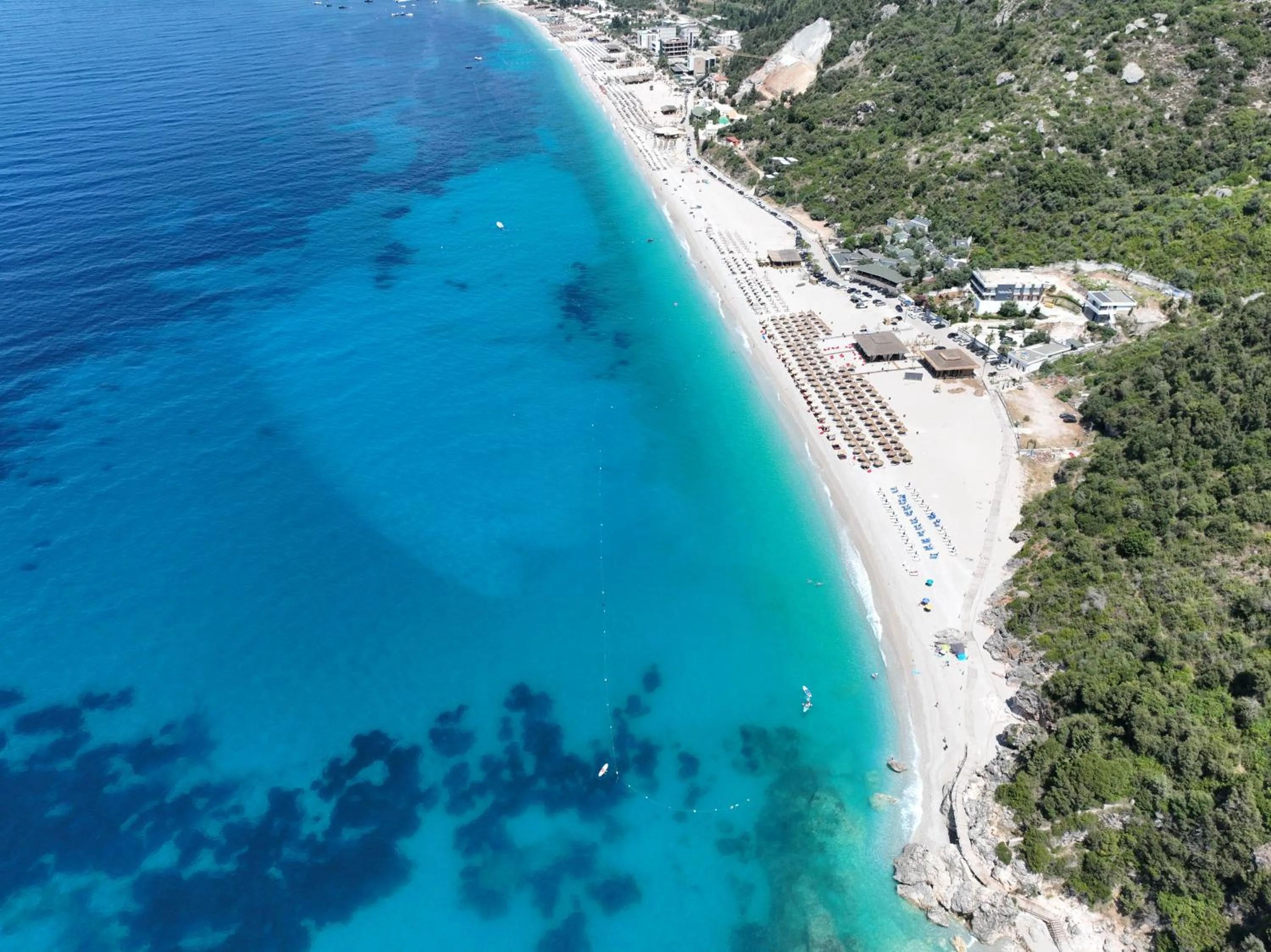 Dhërmi Beach from the air: turquoise water over sand, rows of umbrellas, and the beach ending in rocks and green hillside at its southern end