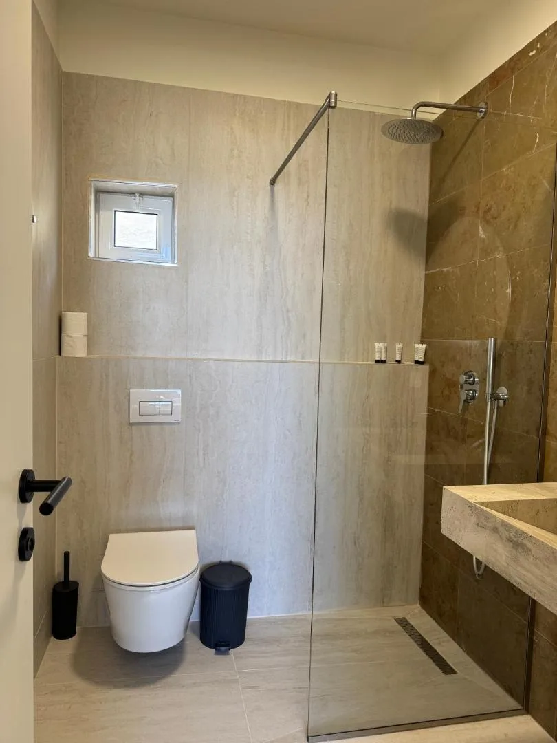 A walk-in shower room in warm stone tiles with a glass screen, rain shower and a wall-hung toilet