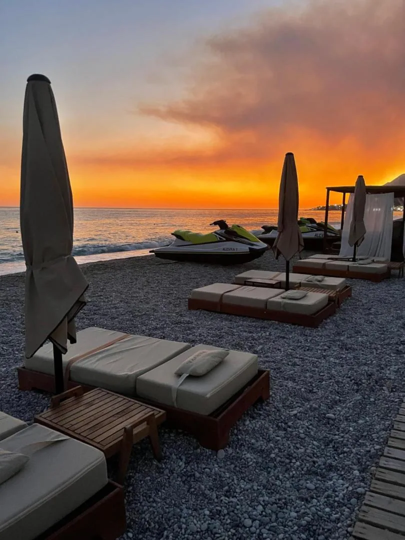 Furled umbrellas and empty loungers on dark shingle under an orange sunset sky
