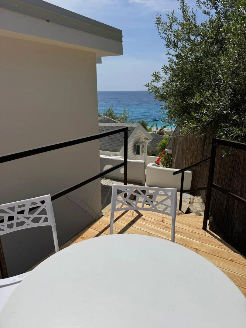 A timber terrace with a round table and white chairs, olive trees and a glimpse of the sea past the rail