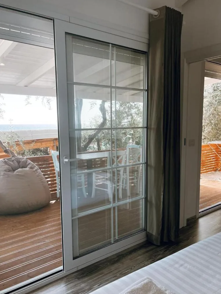 The view from a bungalow bed through glass doors to a timber veranda with a beanbag, olive trees and the sea