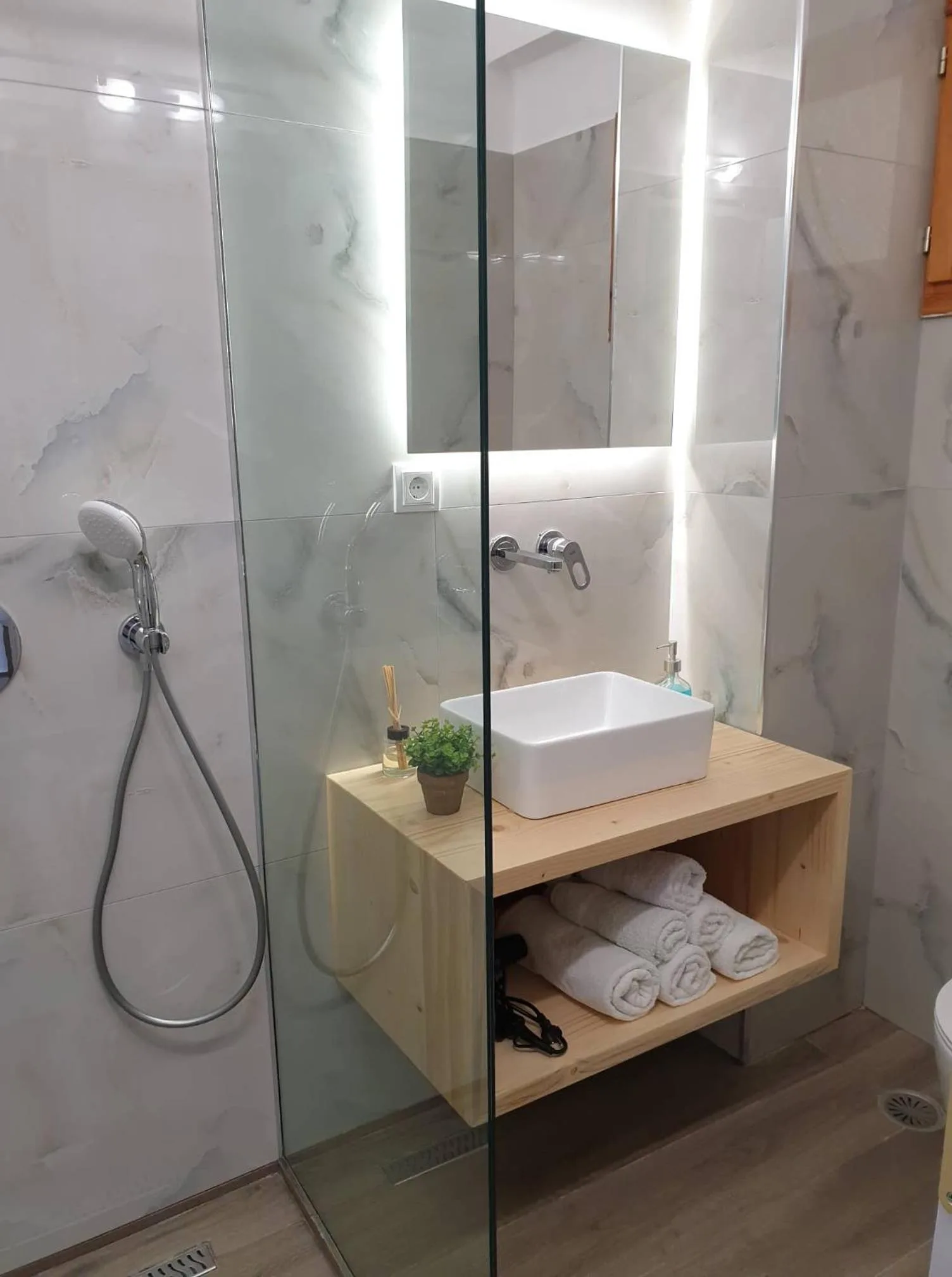 A shower room in white marble-effect tile with a glass screen, a vessel basin on a pine shelf and a lit mirror