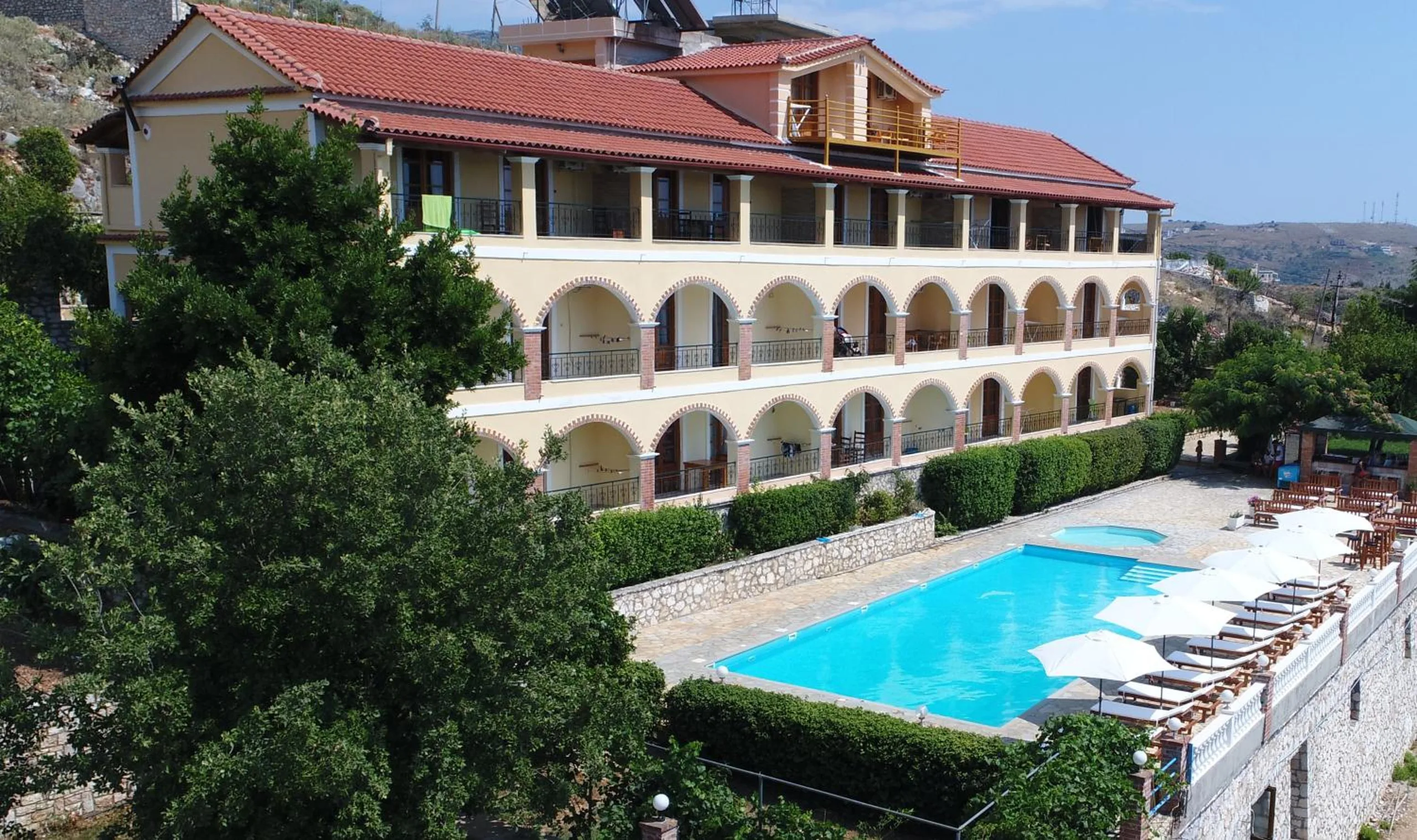 The long cream building with two storeys of brick-trimmed arches above the pool and a row of white umbrellas