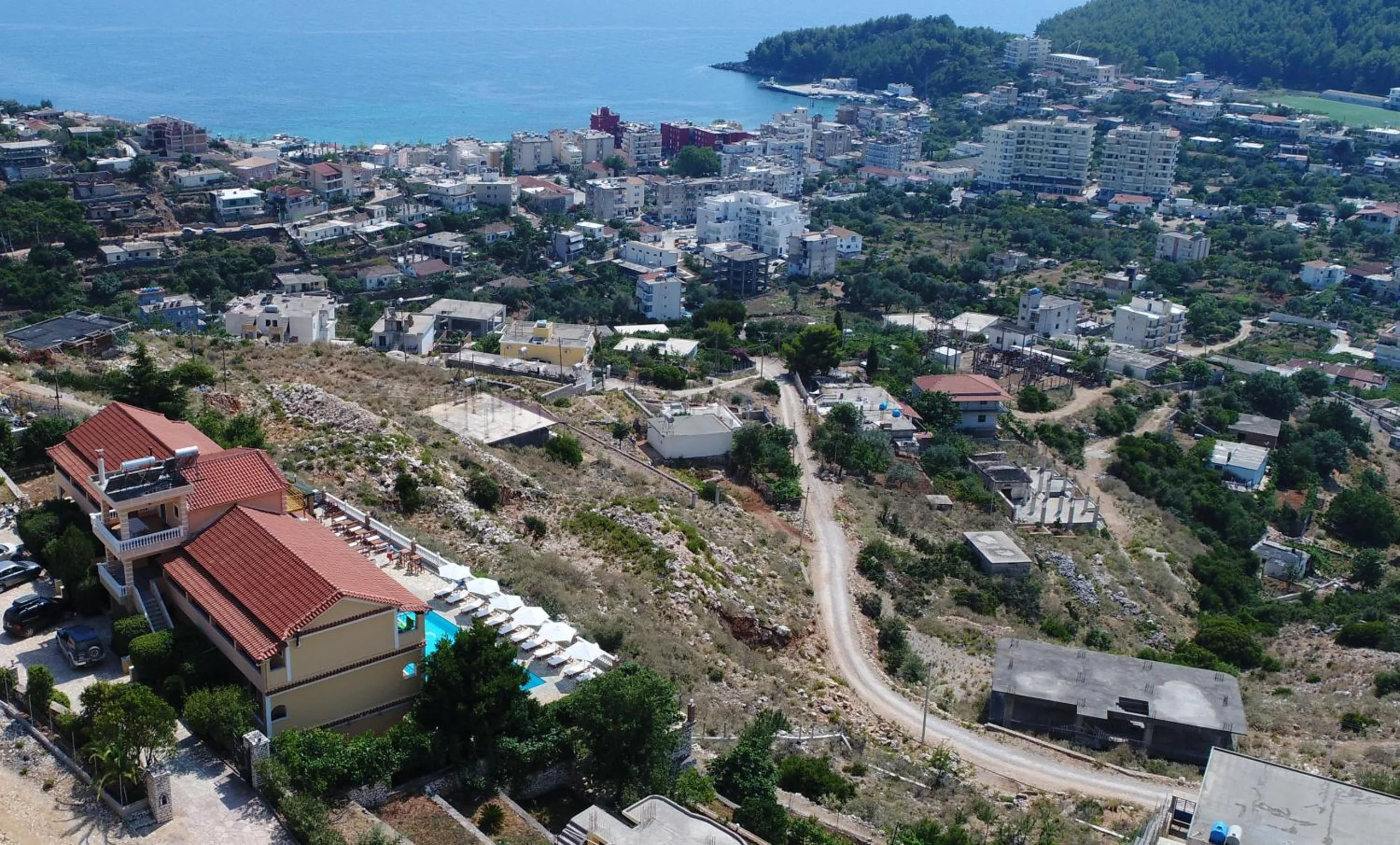 Aerial view of the hotel's red tile roofs and pool on the hillside, with Himarë town and the blue bay below