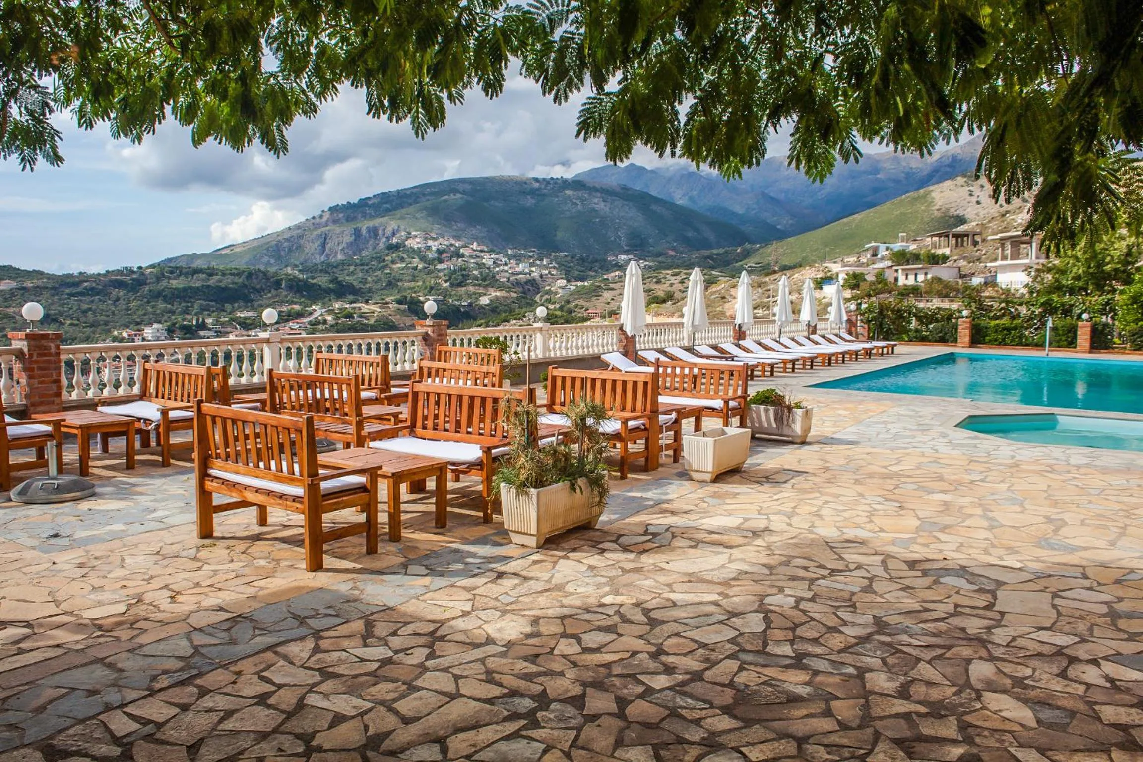 Wooden tables and chairs in the shade of a tree on the flagstone terrace, the pool and mountains behind