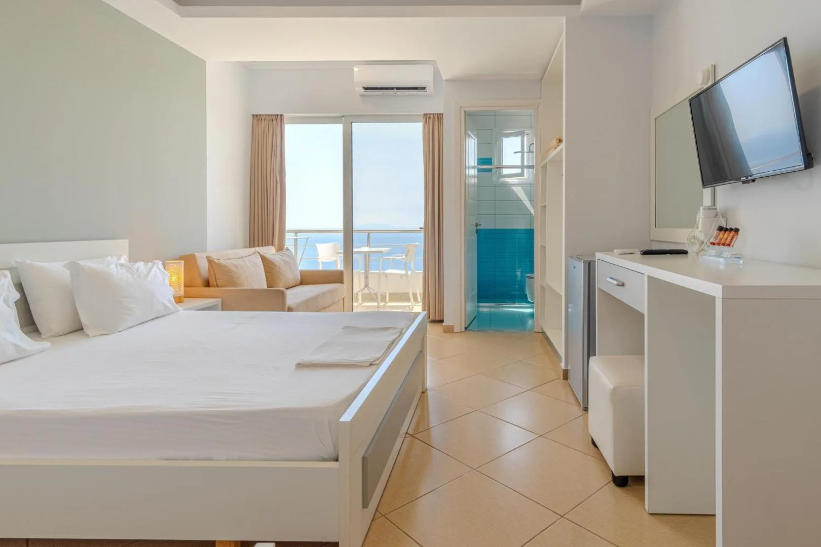 A white room with a double bed and a sliding glazed door onto a balcony, the sea in the opening