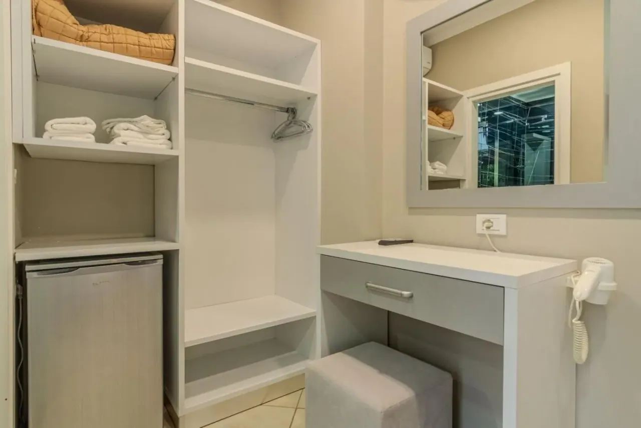 An open wardrobe with folded towels beside a white desk, a mirror, a hairdryer and a small refrigerator