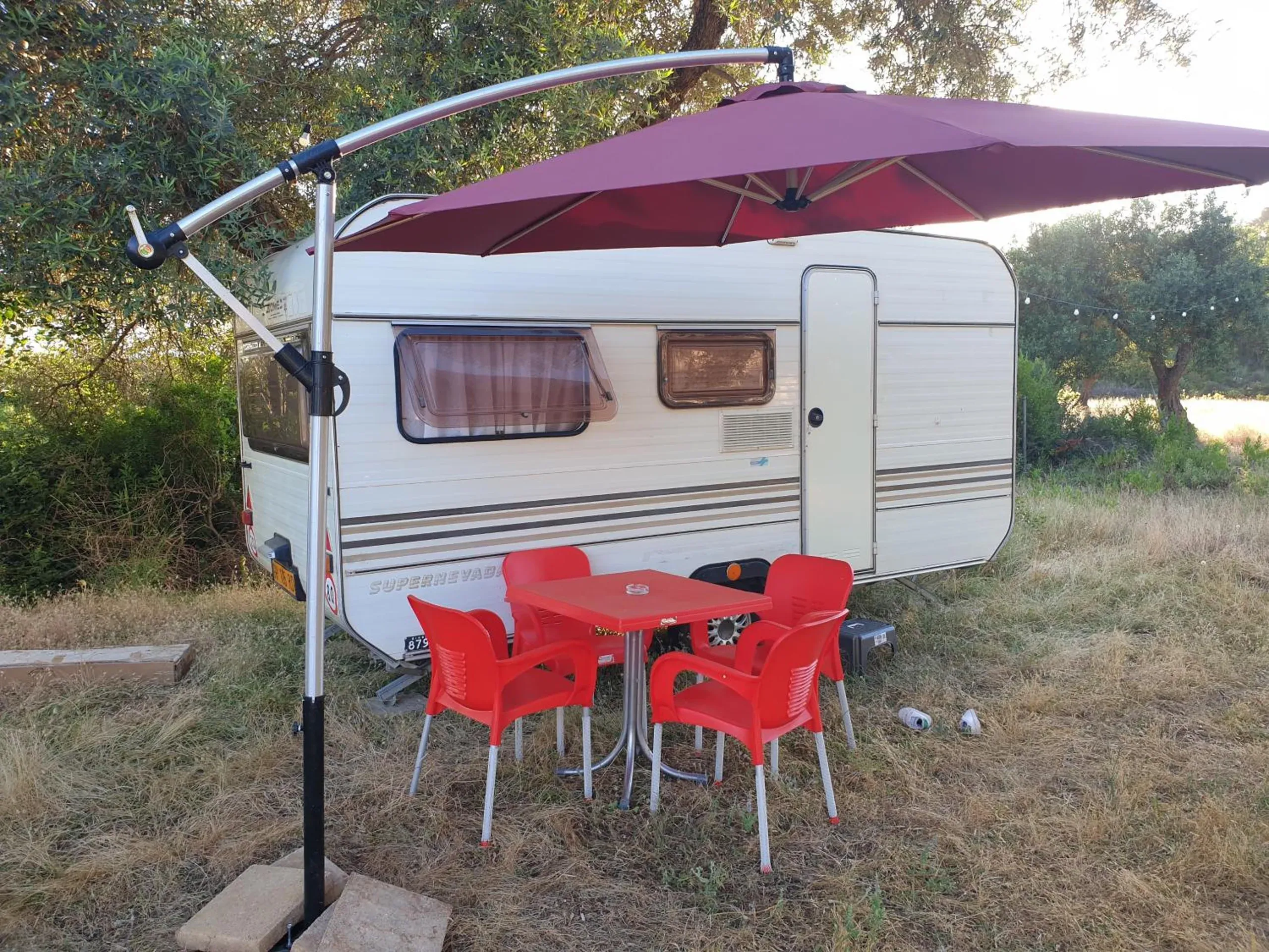 A white caravan with brown livery stripes under a wine-coloured parasol, a red table and chairs in the grass in front of it