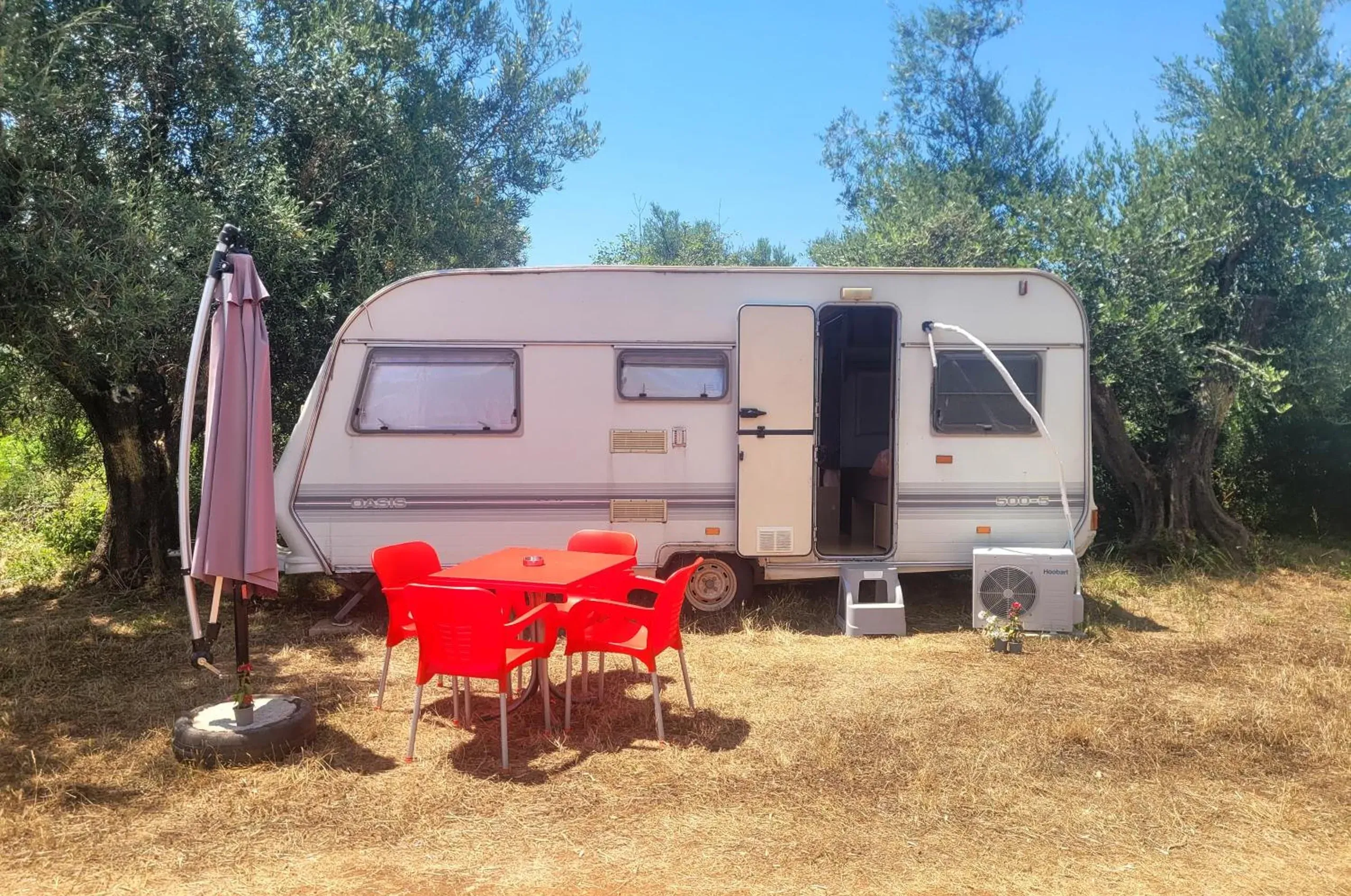 A caravan between olive trees with its door open, an air-conditioning unit beside the step and a red table set out in front