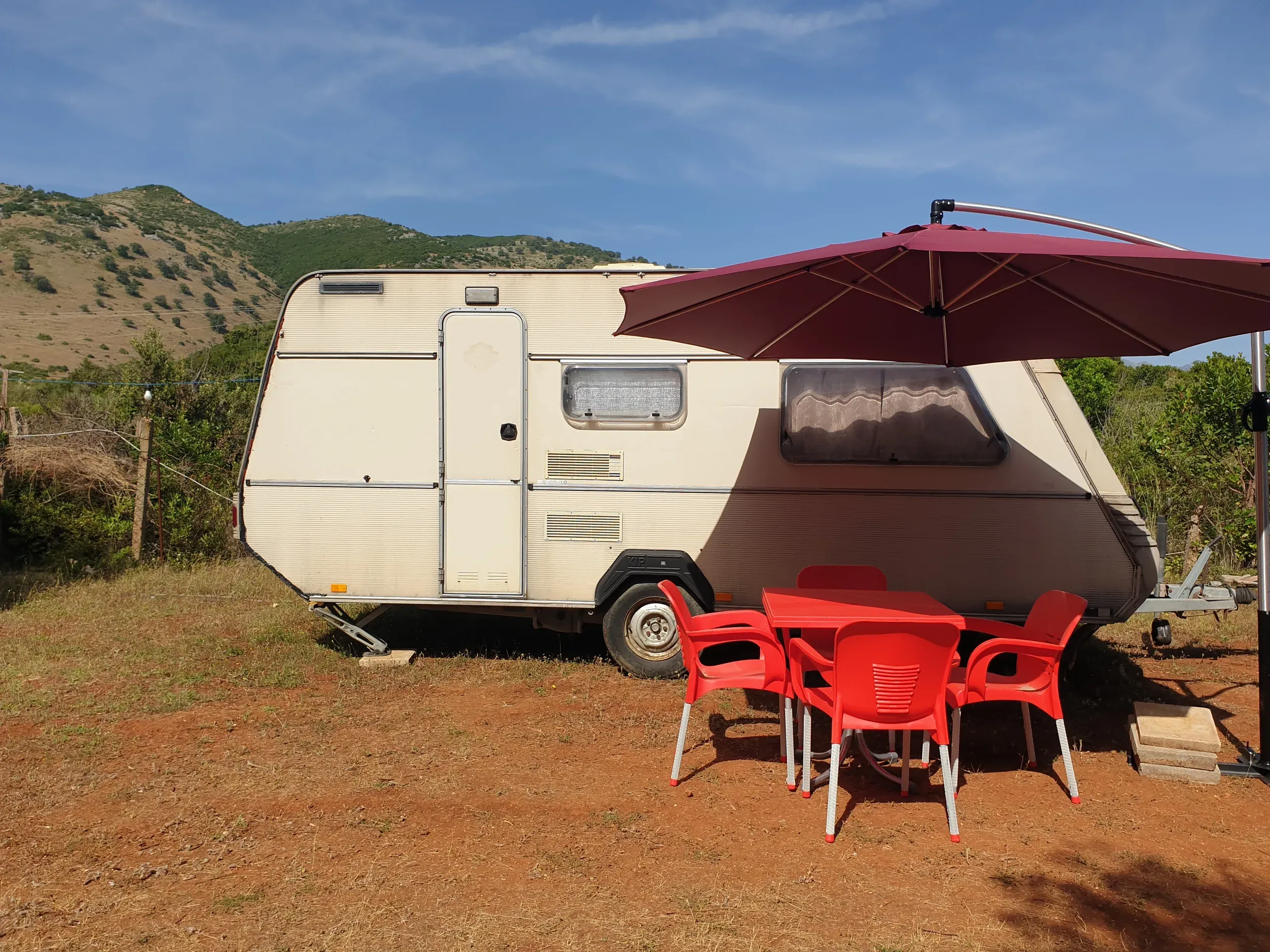 A cream touring caravan on red earth with a wine-coloured cantilever parasol over a red table and four red chairs, green hills behind