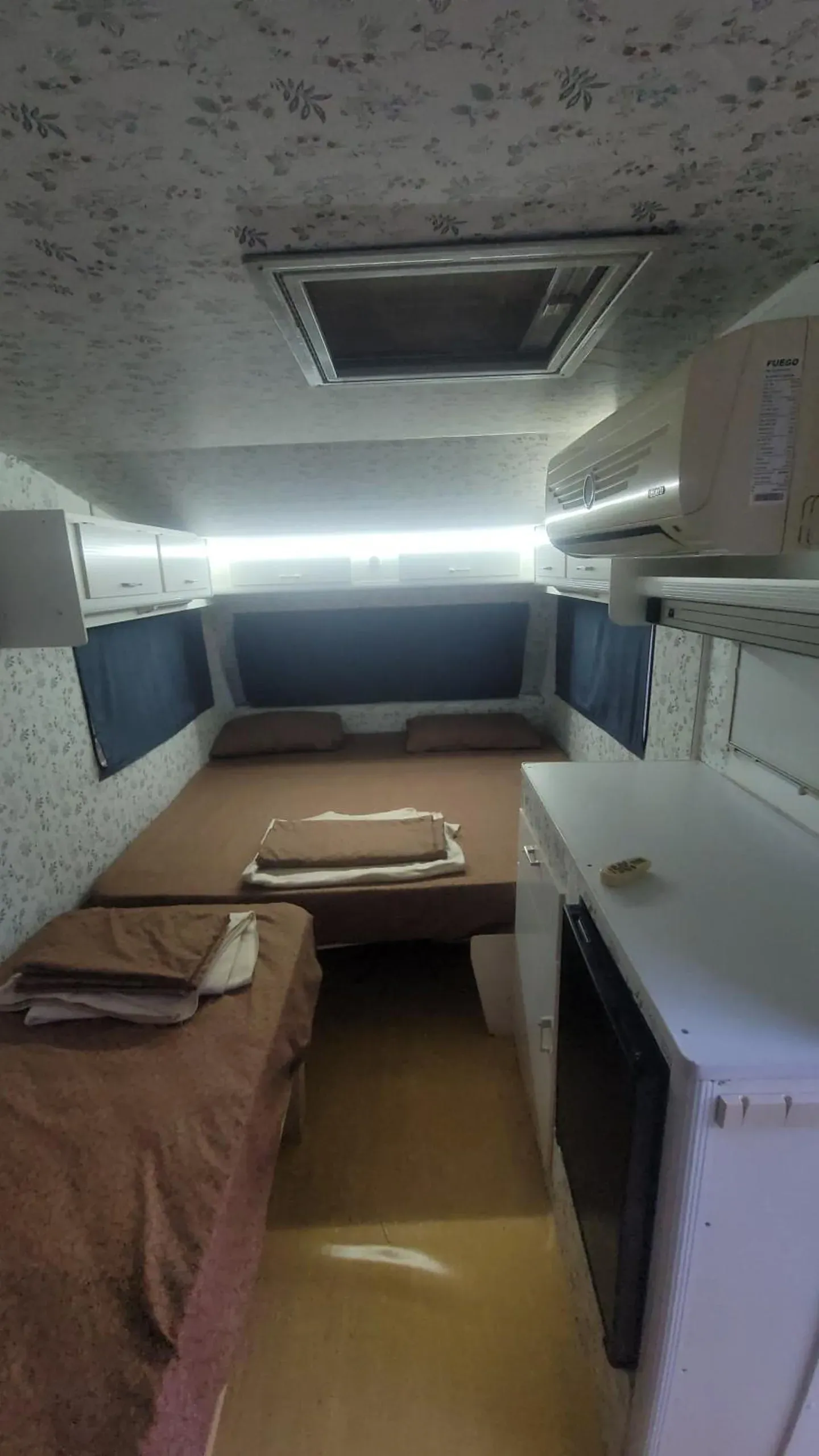 Inside a caravan: a double bed across the end with folded towels and sheets, blinds over the windows and air conditioning above