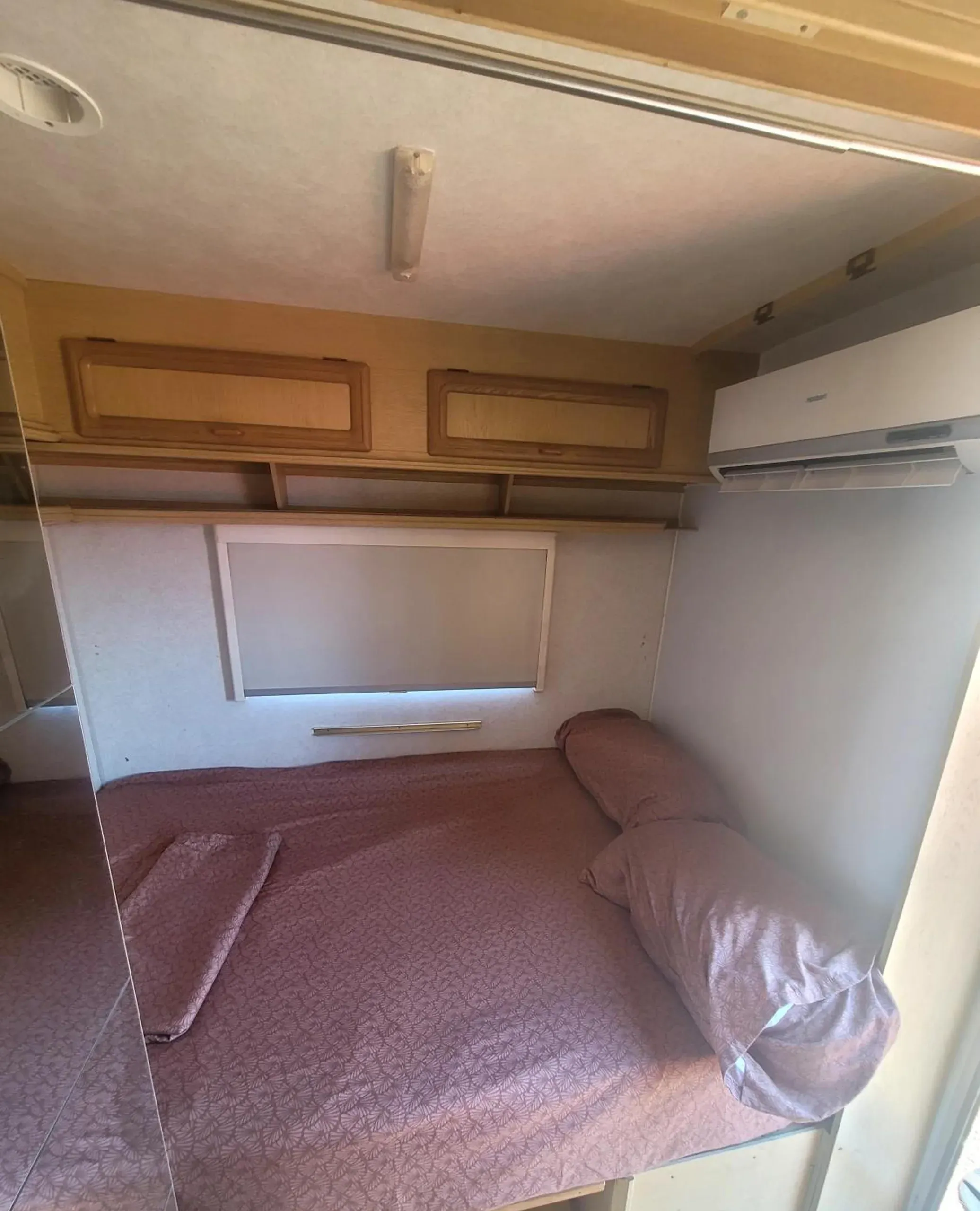 A double bed with rose-coloured covers at the end of a caravan, a wall-mounted air-conditioning unit and timber lockers above