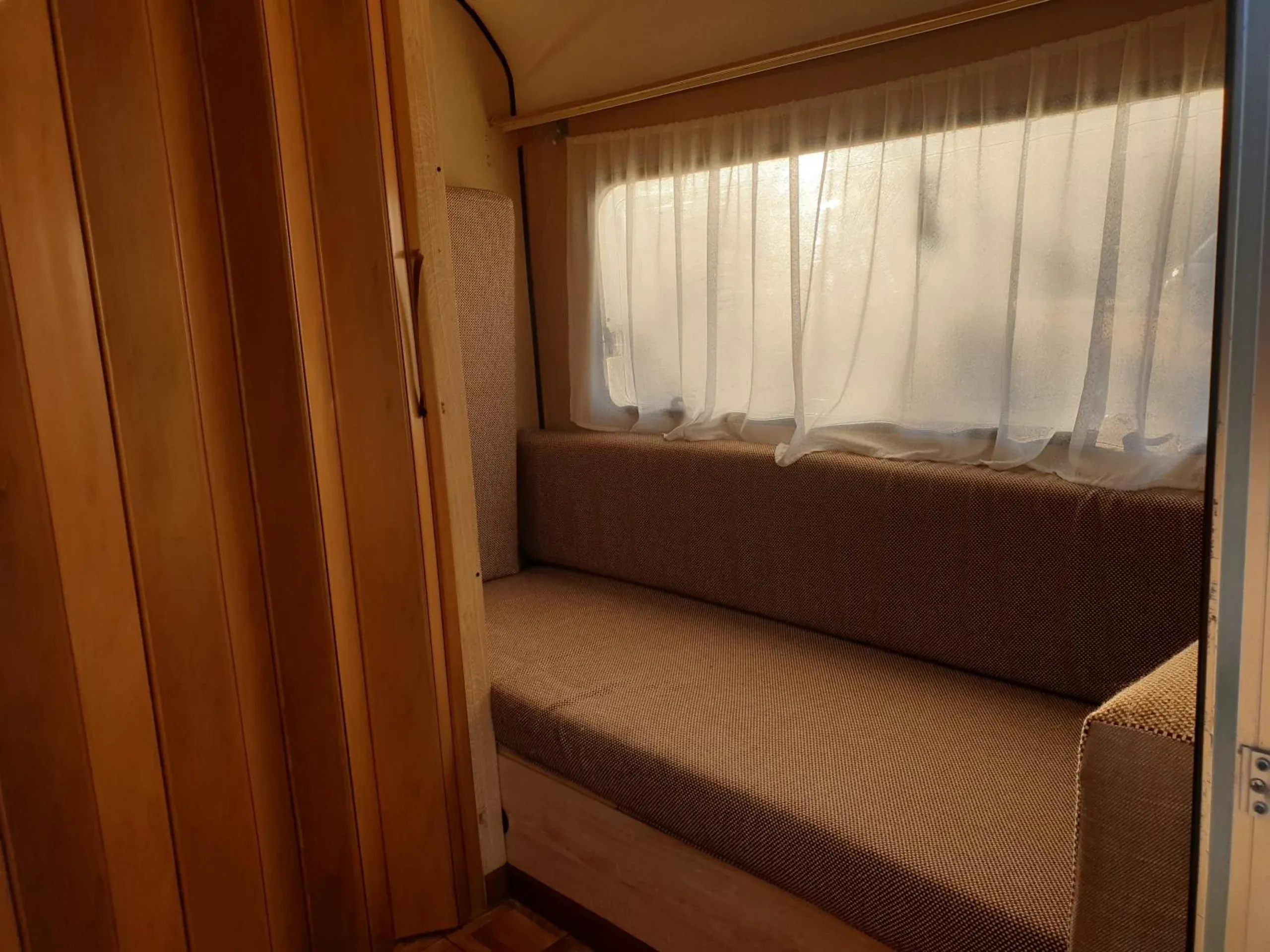 A cushioned bench seat under a net-curtained window inside a caravan, timber panelling on the left