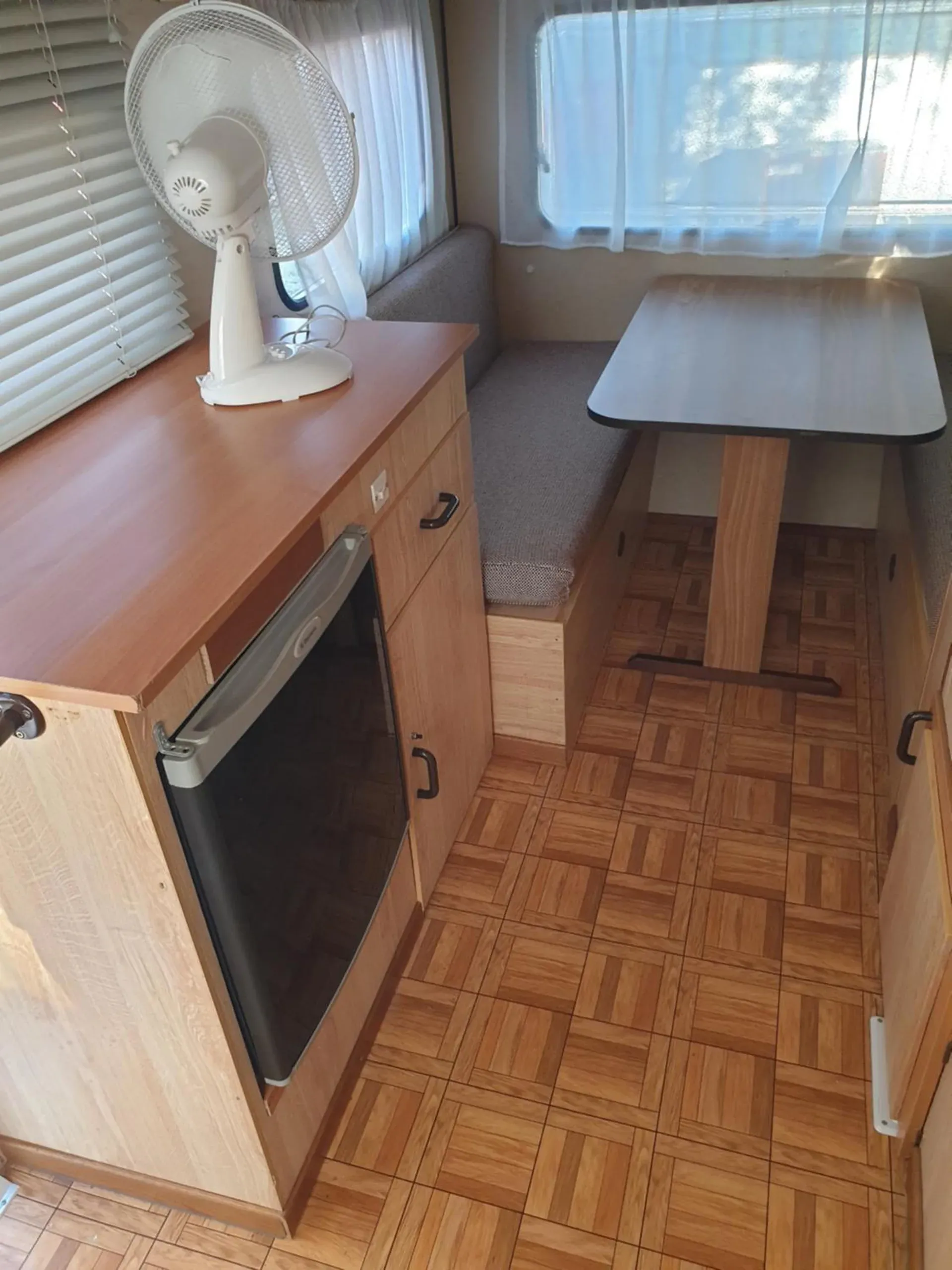 A caravan galley with timber cupboards, a small table on a single leg, a bench seat and a fan, on a parquet floor