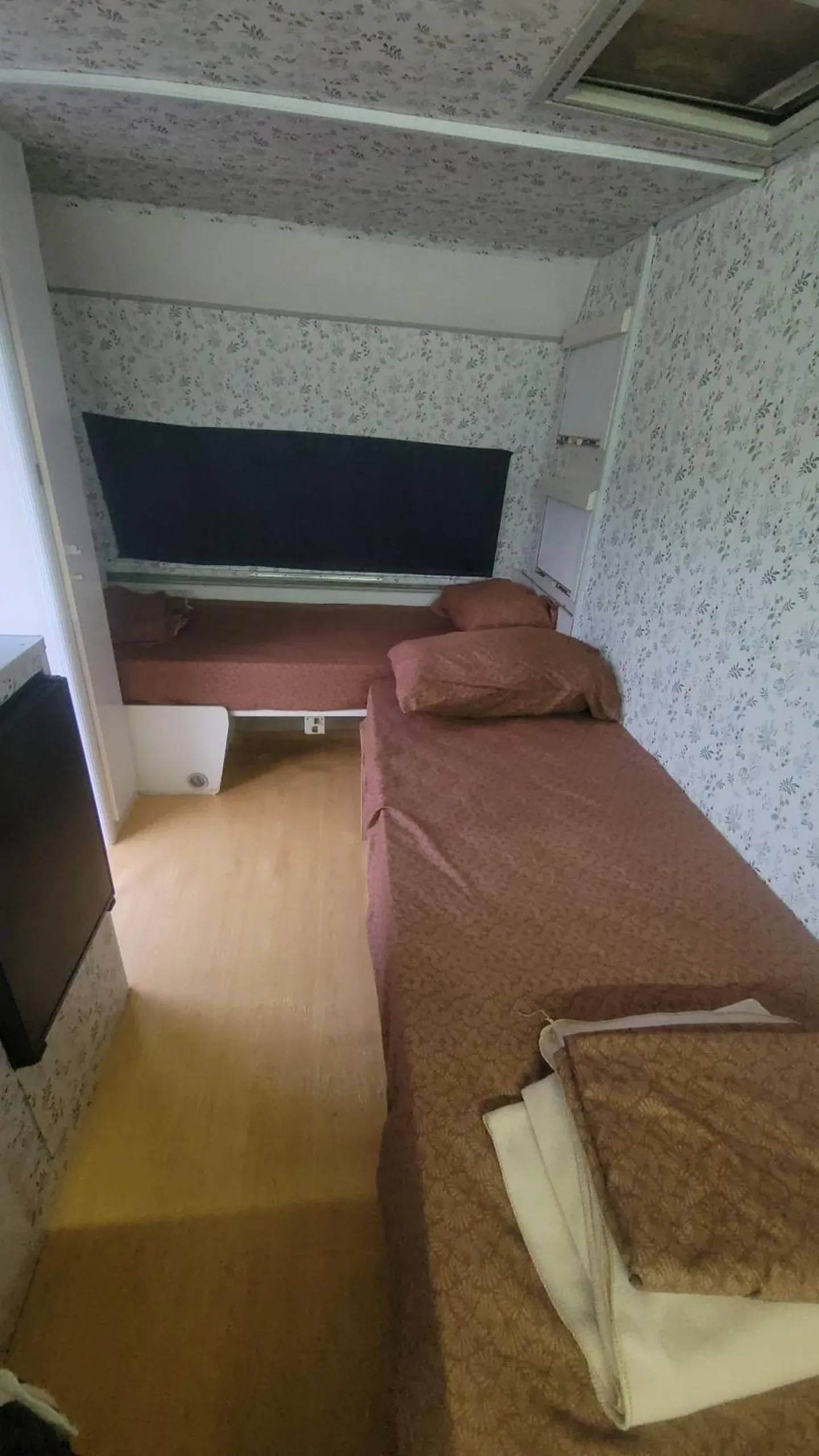 Two beds at right angles in a caravan with floral wallpaper, rose-coloured covers and a pale wood floor
