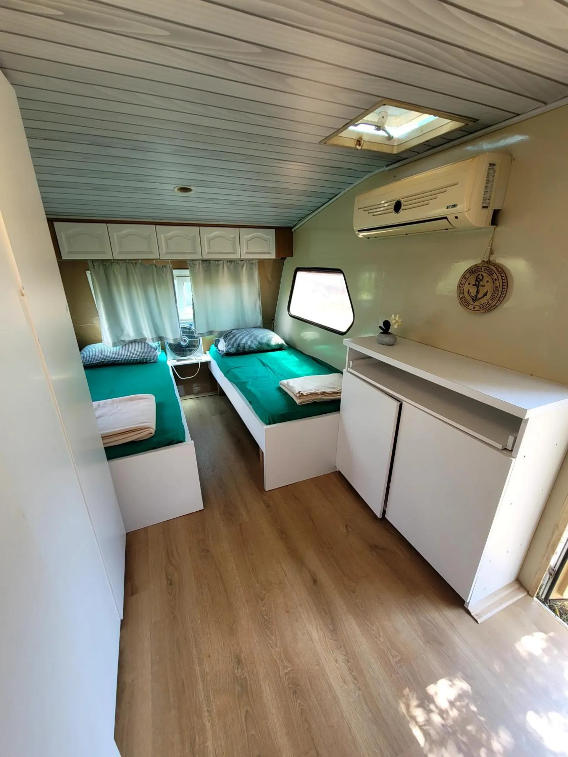 Inside a caravan: two single beds with green sheets either side of a little table, net curtains, white cupboards and an air-conditioning unit on the wall
