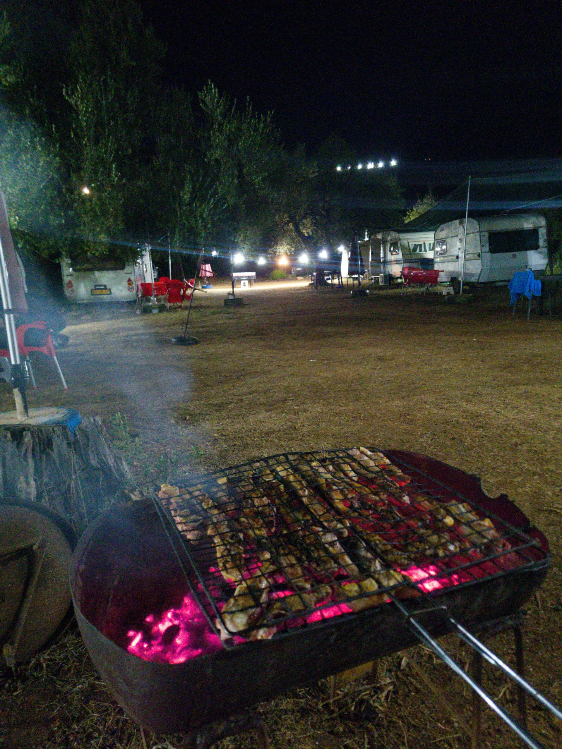 Meat grilling over glowing coals on a round barbecue in the foreground, the lit caravan lane under the olives behind