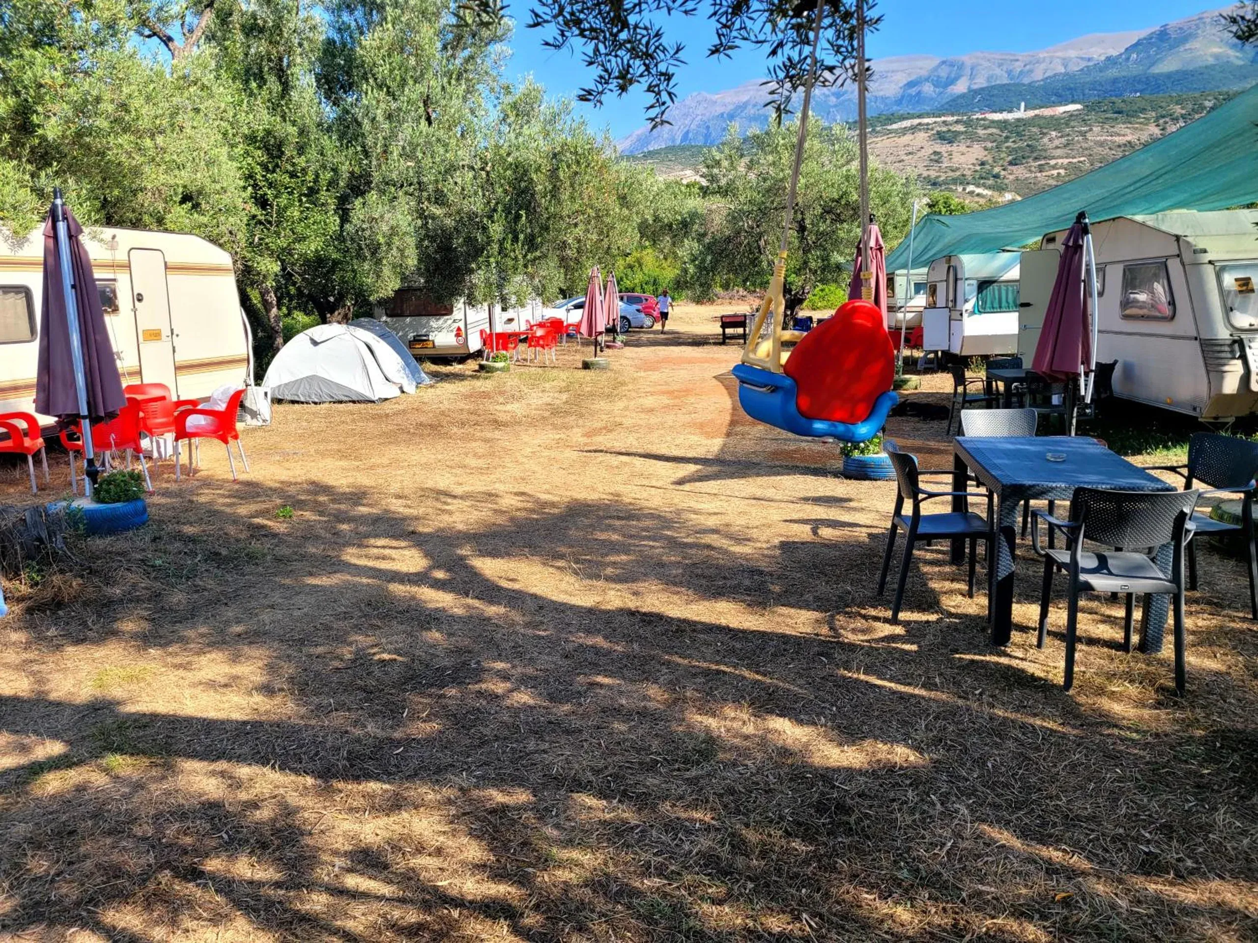 The same lane by day: caravans with parasols and red chairs on the left, a dome tent, teal shade sails on the right and the mountains above Himarë behind