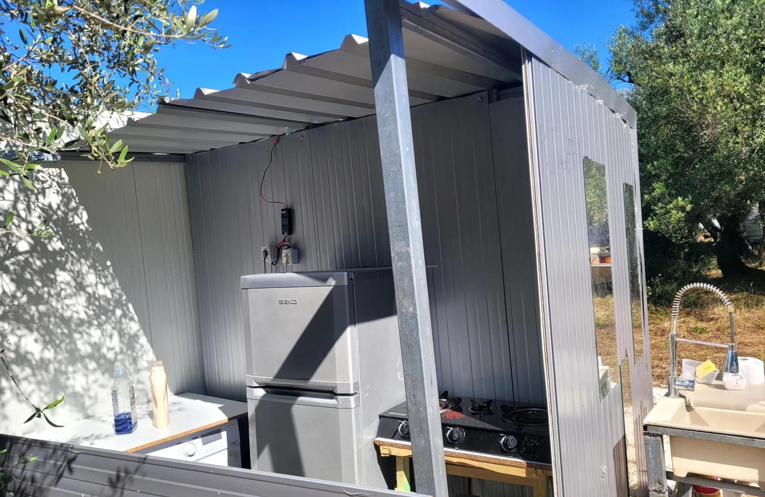 The covered outdoor kitchen: a fridge, a two-ring gas hob and a sink with a spray tap under a corrugated roof, olive trees around it