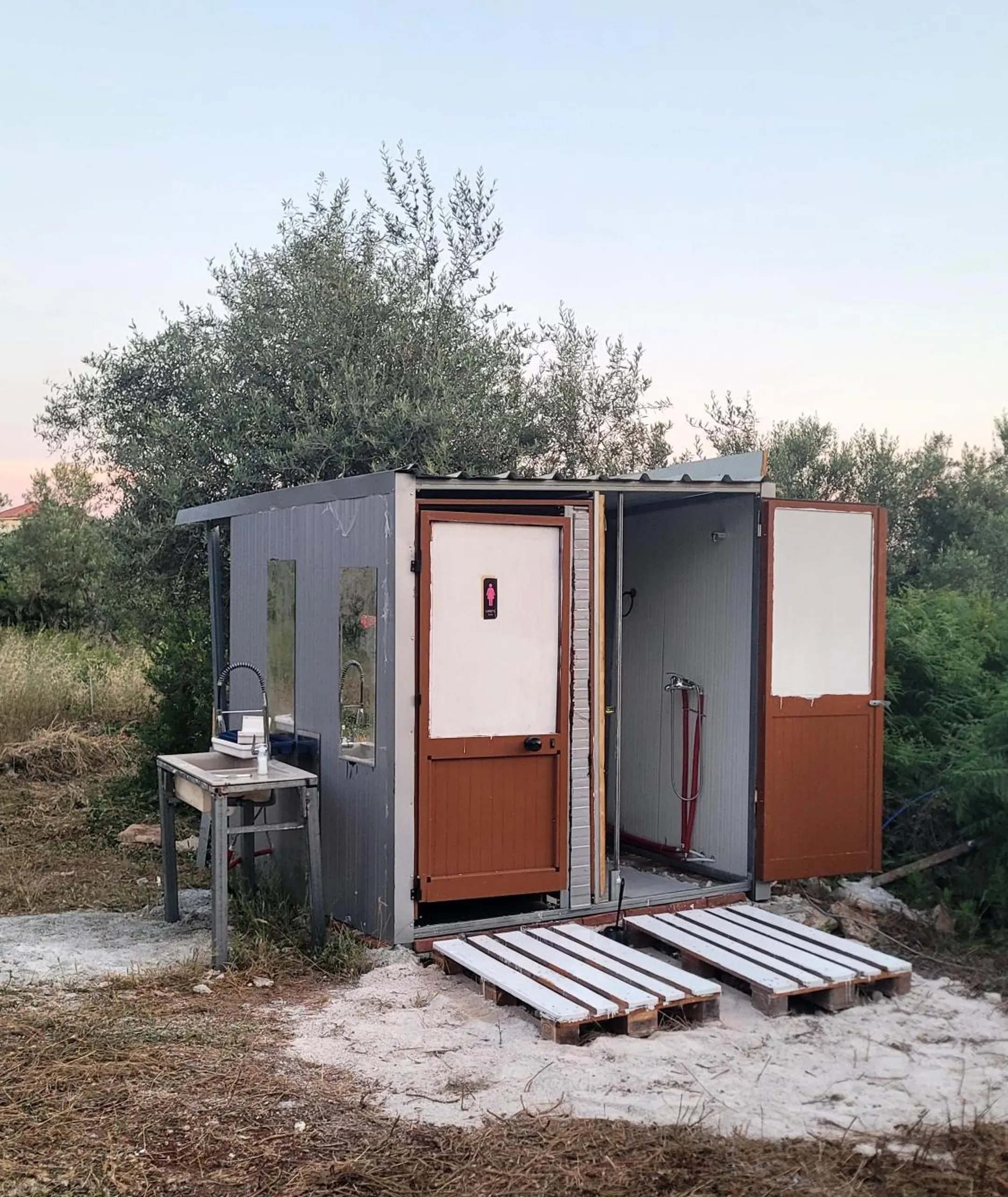 The grey-panel shower and toilet block with brown-and-white doors, standing on white pallets on pale gravel, a washbasin outside and olive trees behind