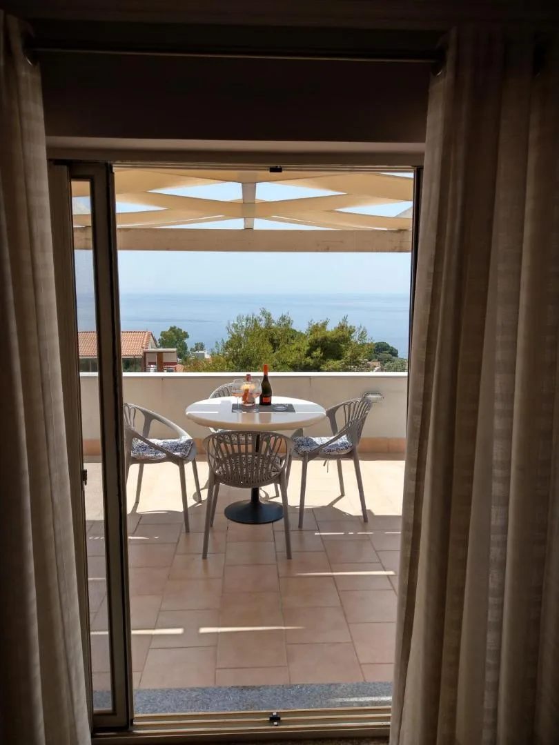 Seen through an open balcony door, a round white table and four chairs on a terrace, with trees and the sea beyond the rail