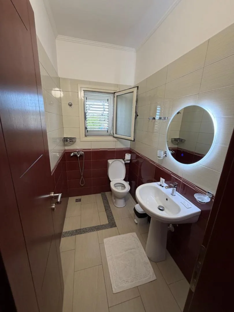A bathroom at Augustus Hotel tiled cream above and claret below, with a shower, a basin under a round lit mirror and a window with white shutters