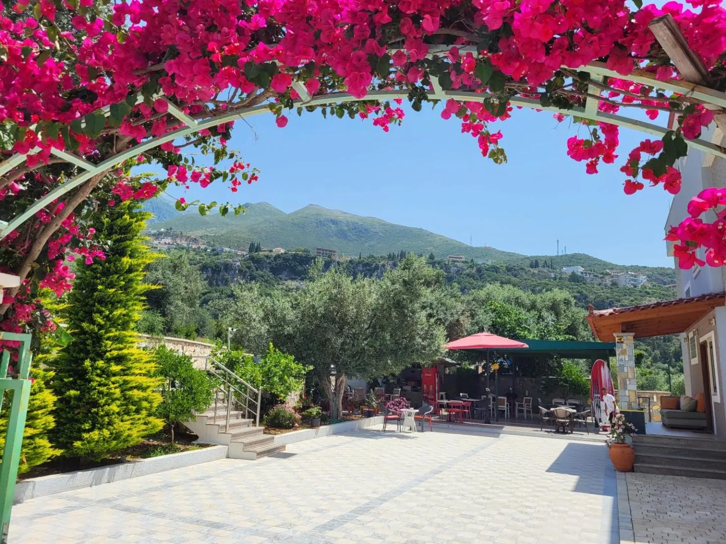 A metal arch thick with magenta bougainvillea framing the courtyard of Augustus Hotel, with cypress trees, an olive grove and the mountains behind