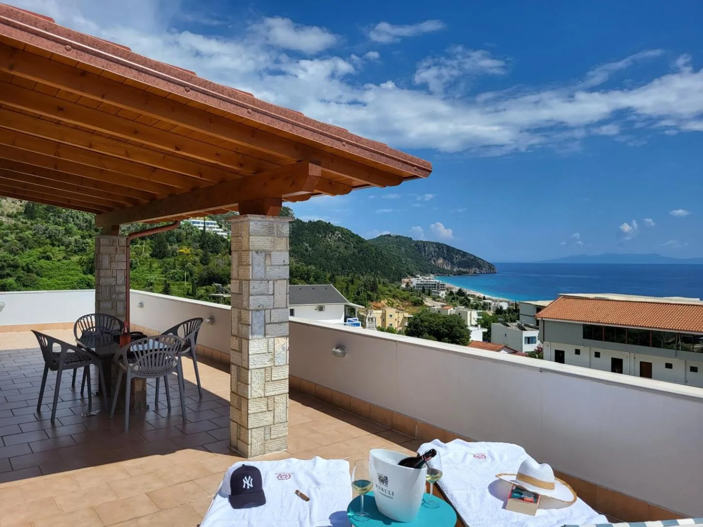 A tiled terrace under a timber roof on stone pillars at Augustus Hotel, with a table and chairs, looking north along the coast to the beach and the hills