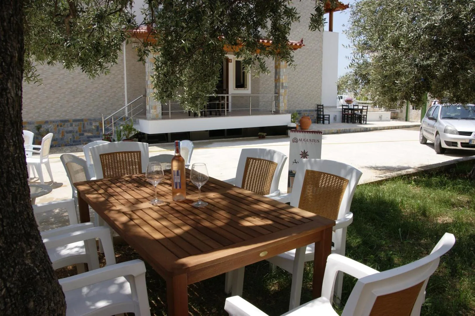 A long wooden table with white chairs in the dappled shade of an olive tree in the garden at Augustus Hotel