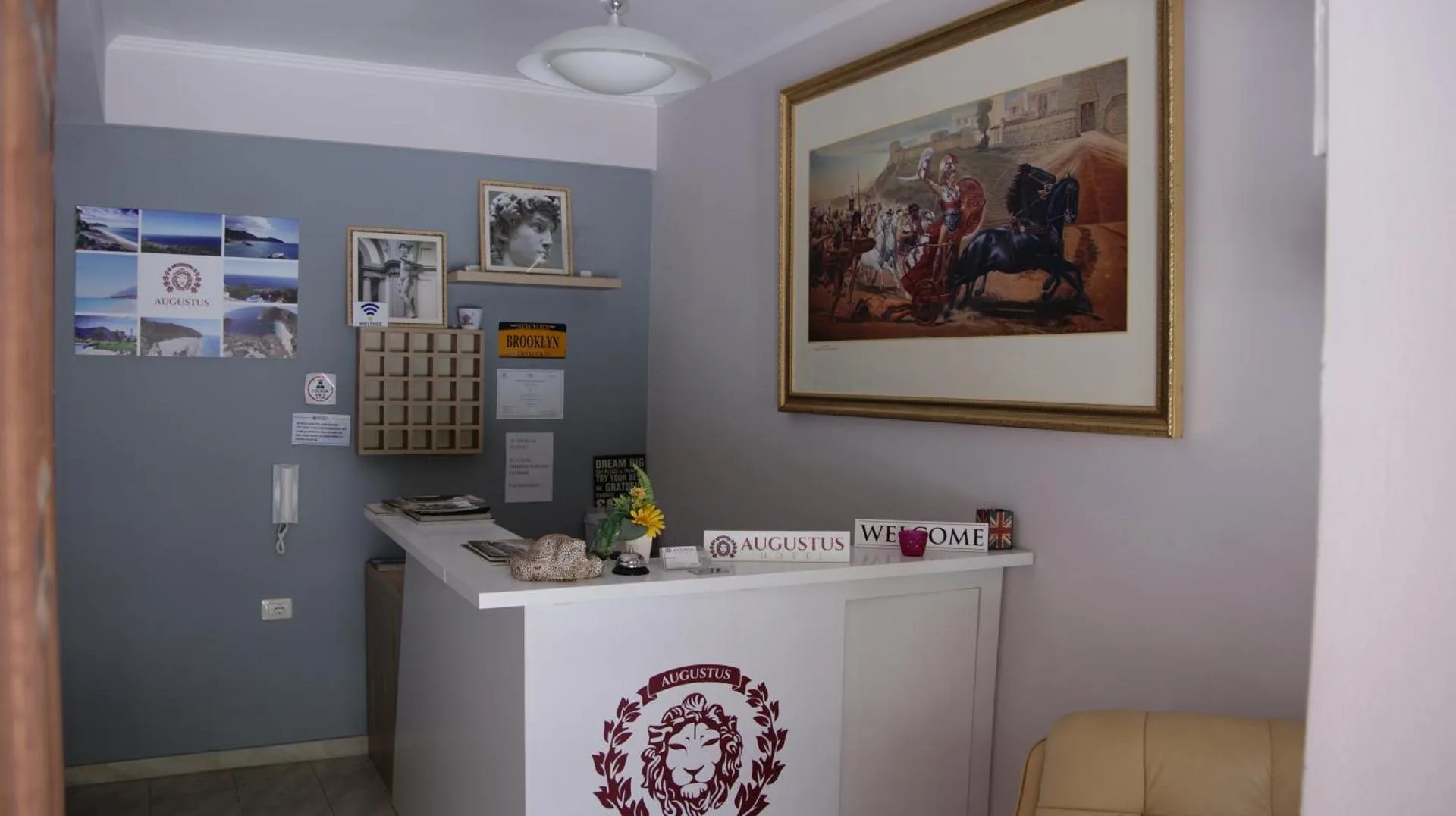 The reception at Augustus Hotel: a white desk printed with the claret lion-in-laurel crest, a framed painting of a chariot battle above it, and photographs of the coast pinned to a grey wall