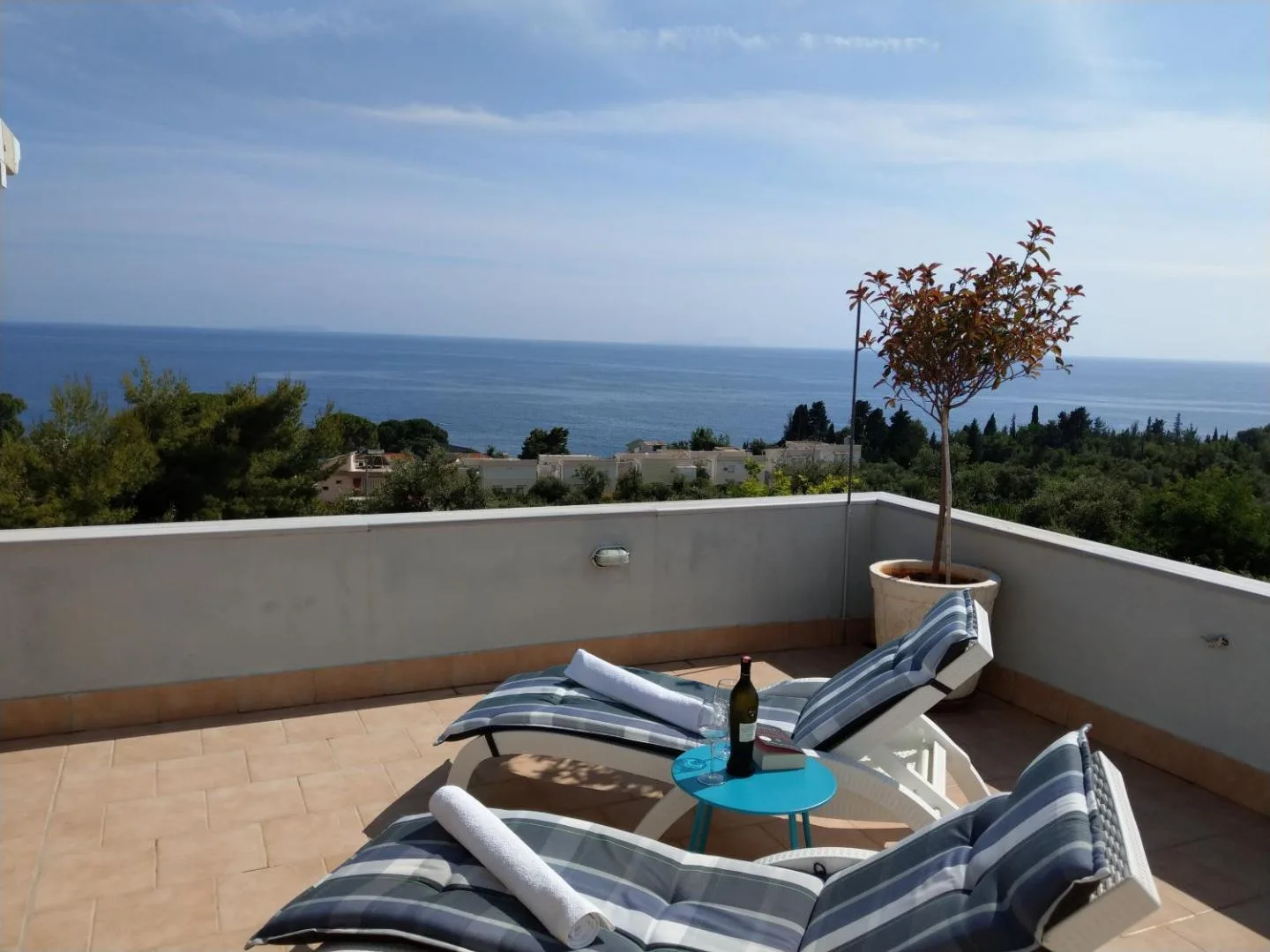 Two padded loungers and a small table with a bottle of wine on a tiled roof terrace, a potted tree at the parapet and the sea along the horizon
