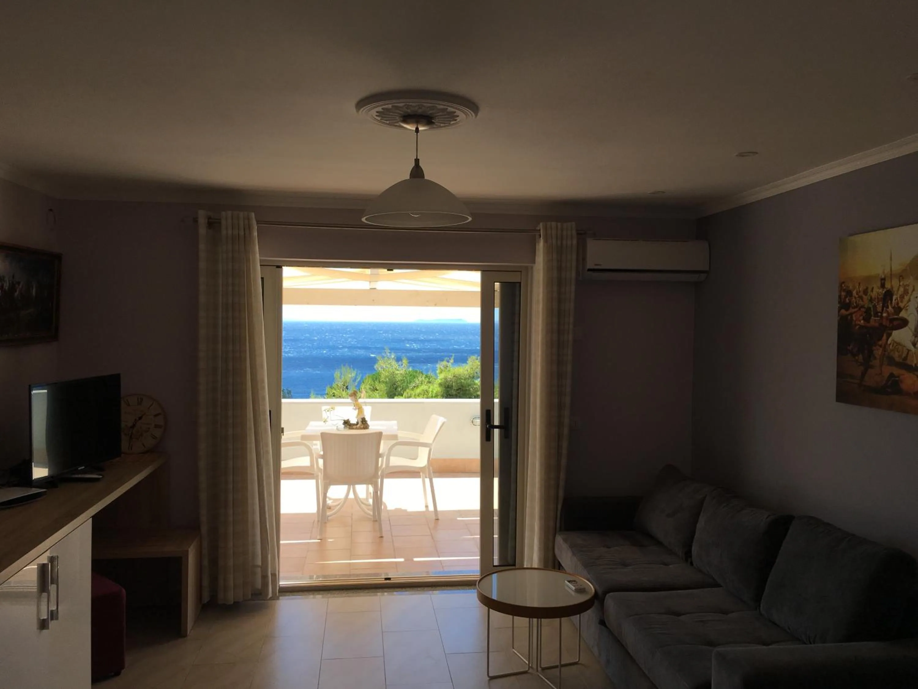 A larger room at Augustus Hotel with a grey sofa, a television on a counter and open glass doors to a terrace with white chairs and the sea beyond