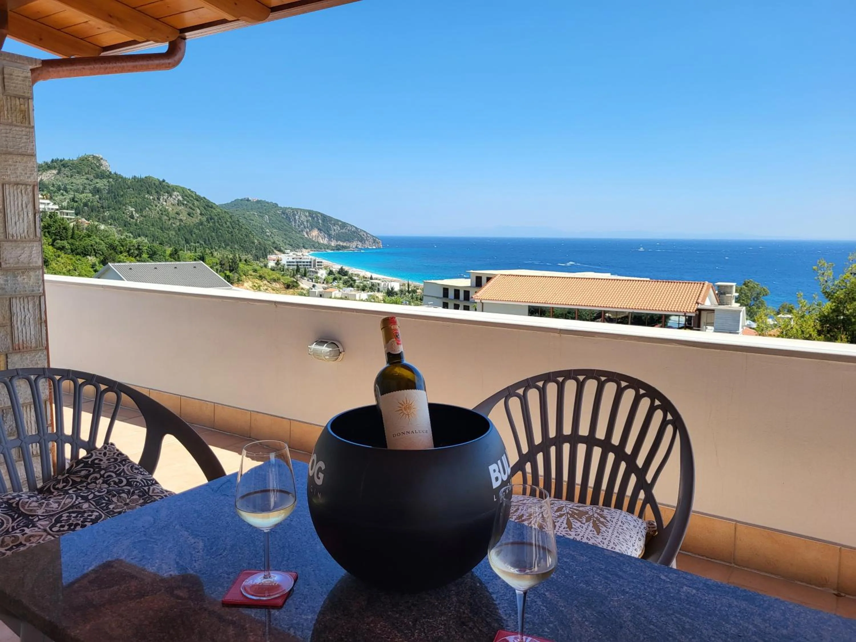 A covered terrace table at Augustus Hotel with a bottle of white wine in an ice bucket and two glasses, looking down over tiled roofs to Dhërmi Beach, the headland and the open sea
